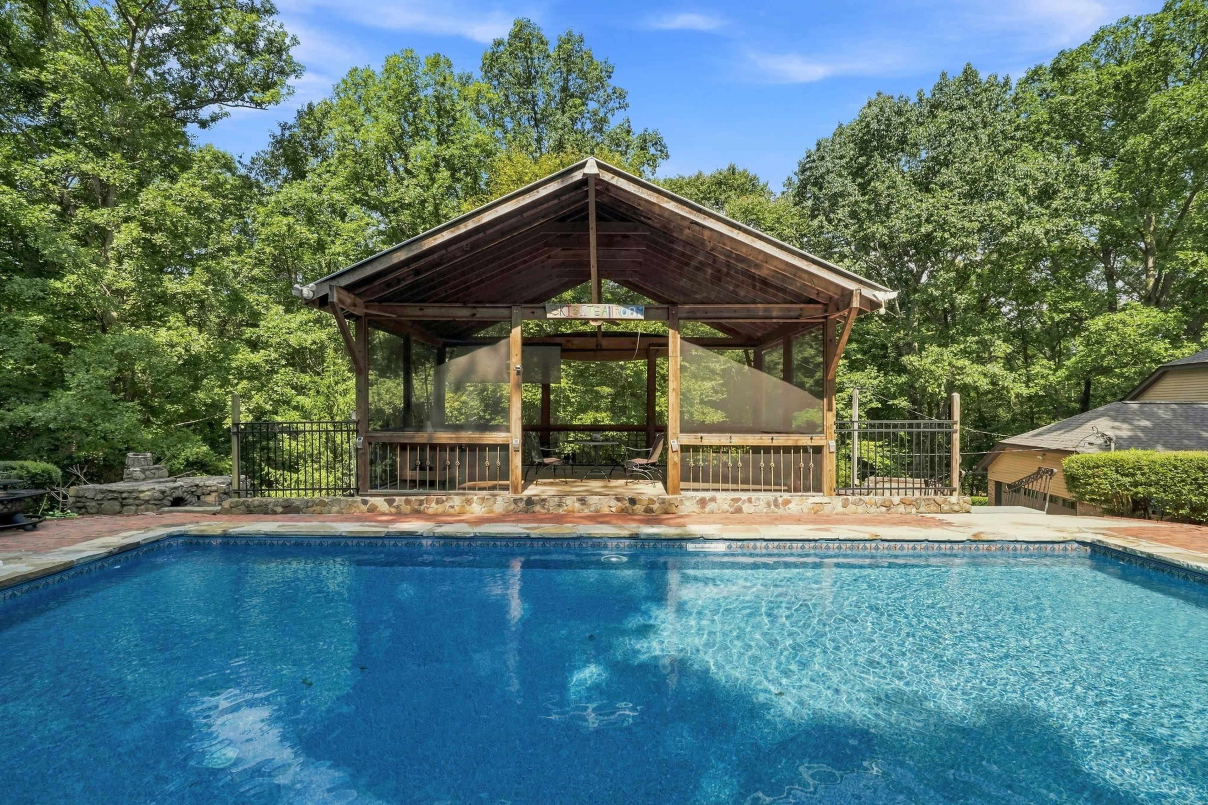 The image shows a pool with a wooden gazebo structure at one end, surrounded by trees.