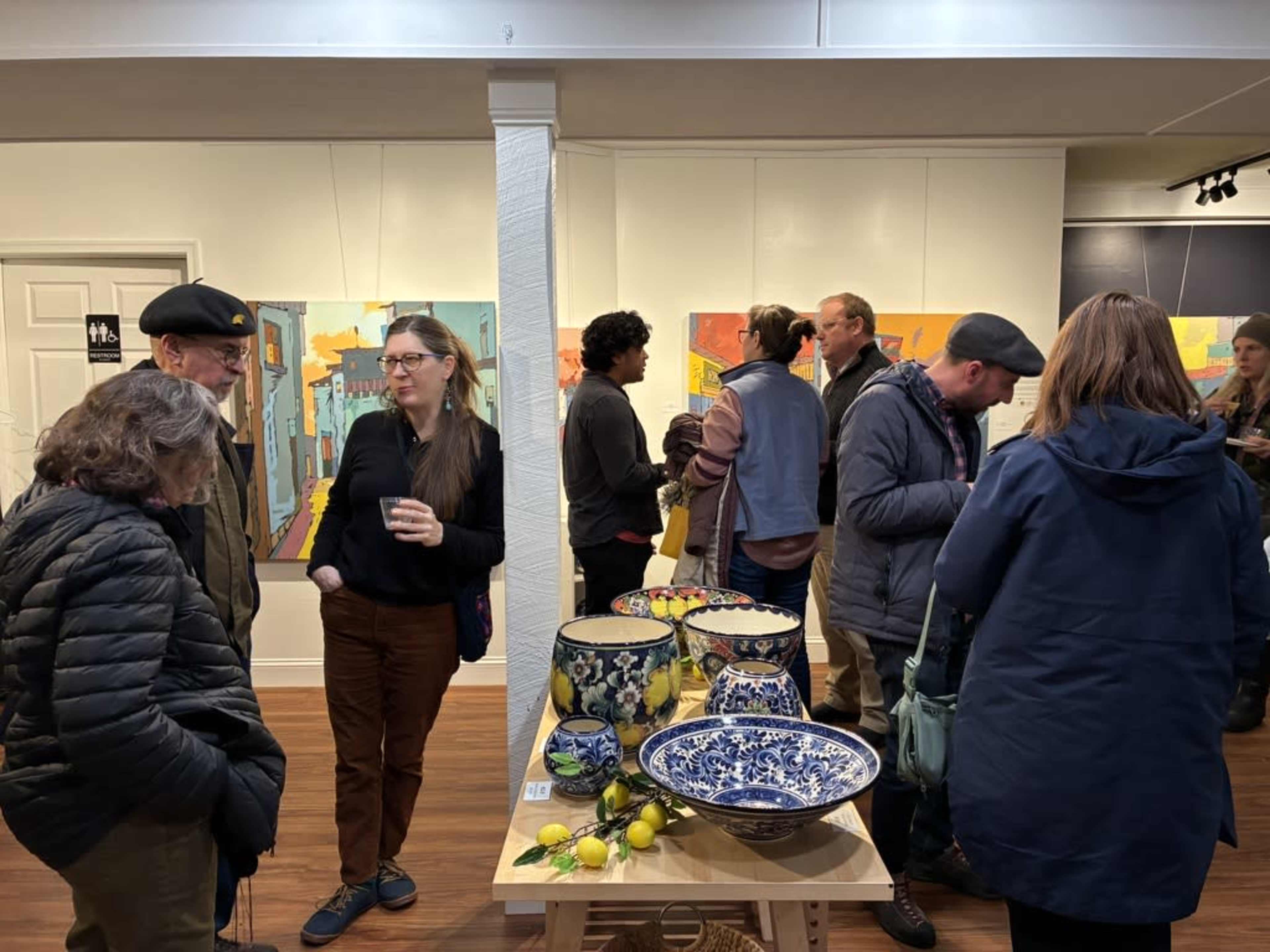 A group of people socialize in an art gallery featuring colorful paintings on the walls and decorative bowls on a table.