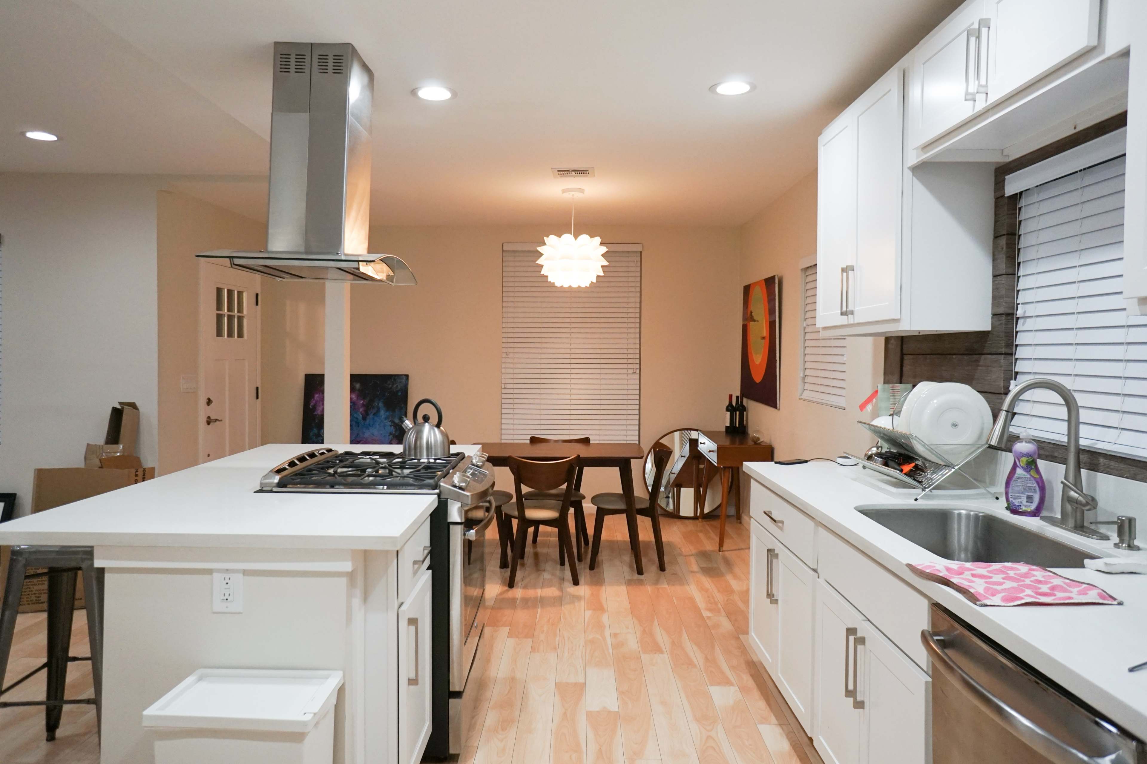 A modern kitchen with white cabinets, a stainless steel range hood, and a wooden dining table adjacent to the space.