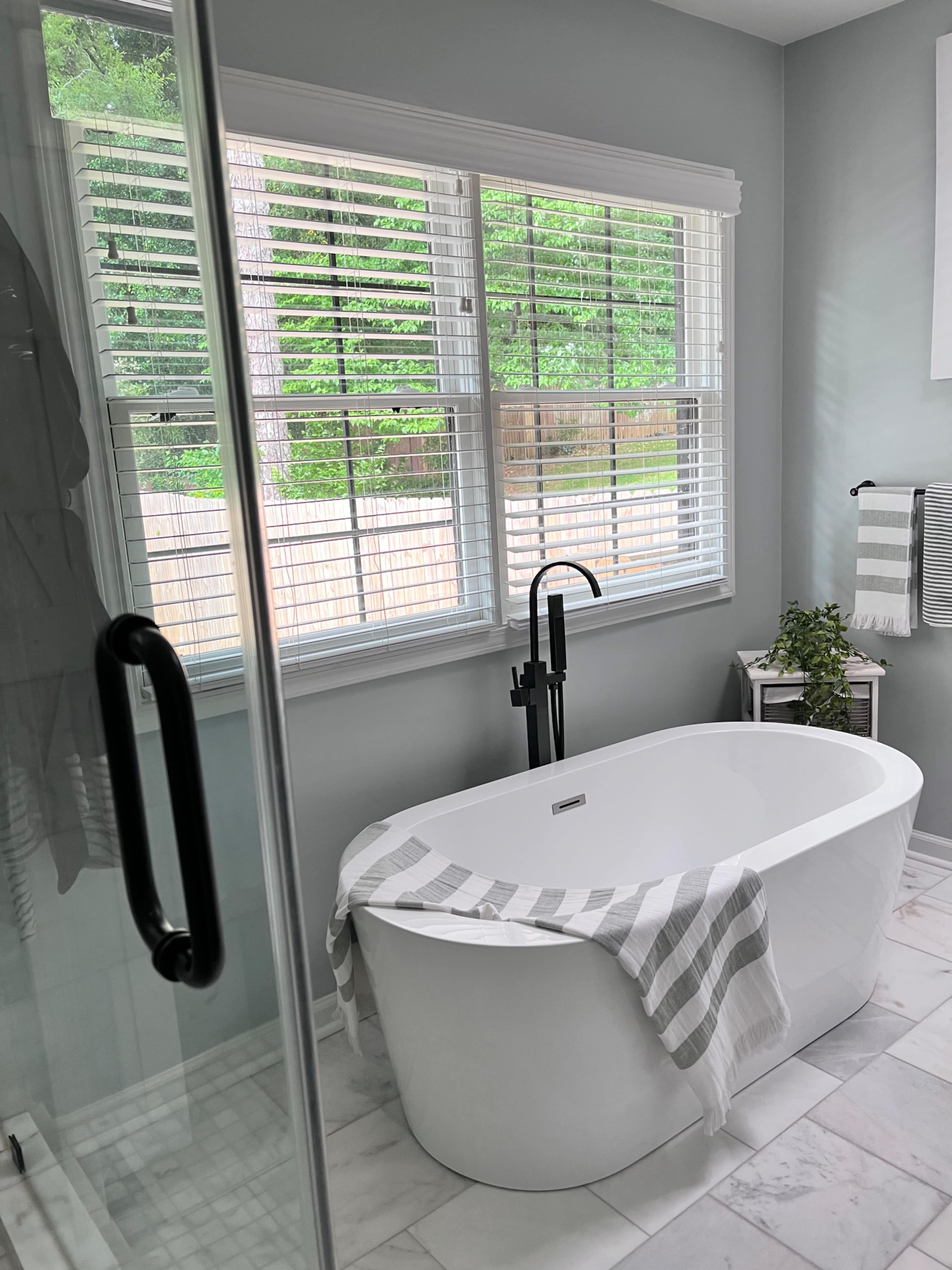 The image shows a modern bathroom featuring a freestanding white bathtub, a glass shower enclosure, and a large window with white blinds.