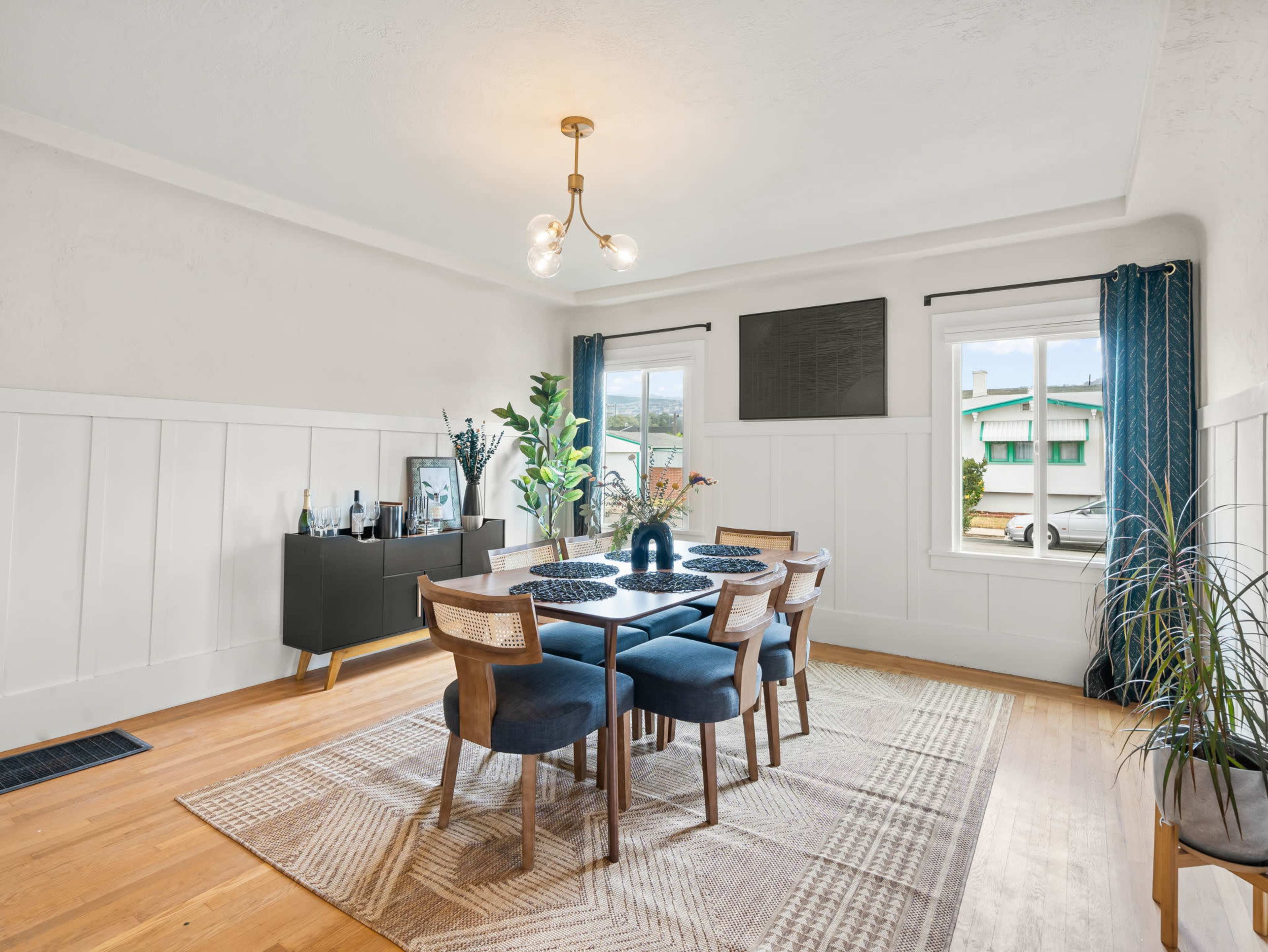 A dining room features a table with four chairs, a sideboard, and two large windows draped with curtains.