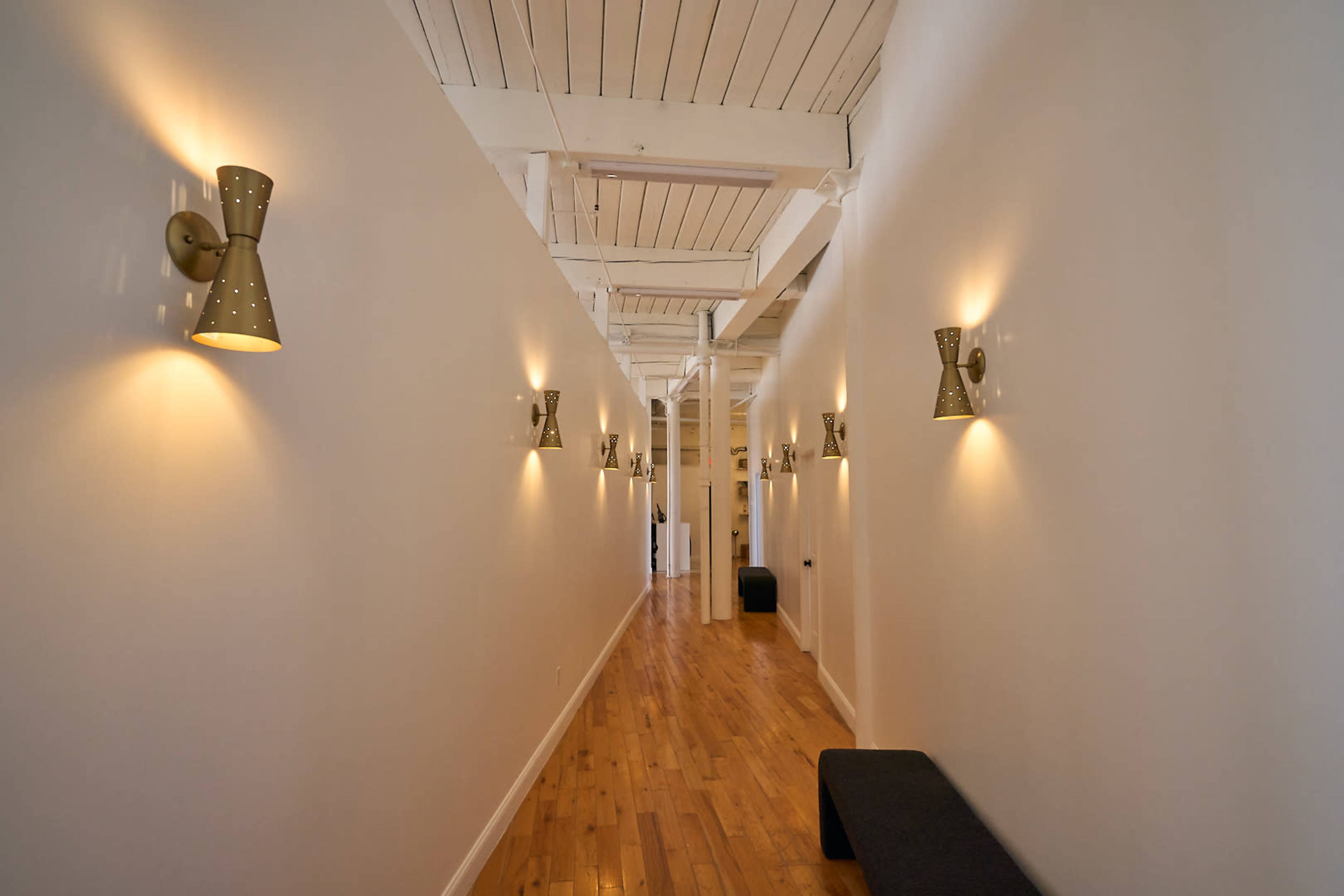 A narrow hallway with wood flooring, white walls, and wall-mounted light fixtures casting a warm glow.