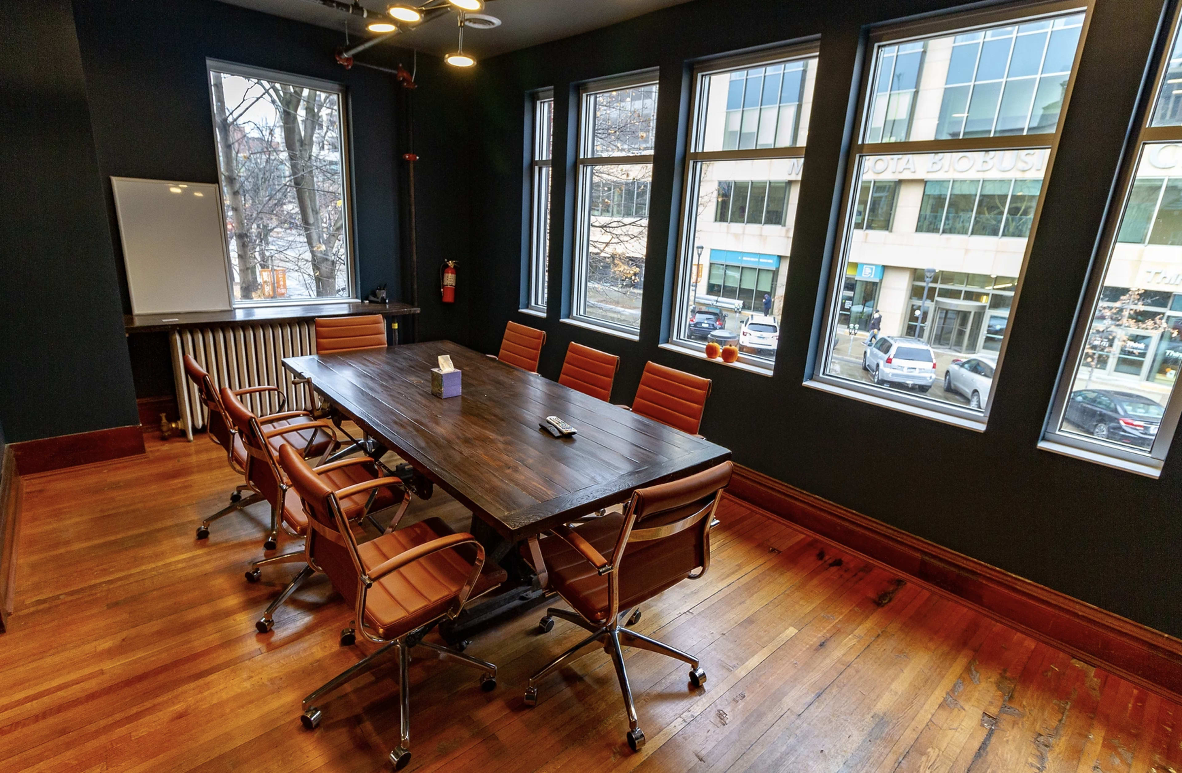A conference room features a large wooden table surrounded by eight orange leather chairs, with large windows allowing natural light and a view of the street outside.