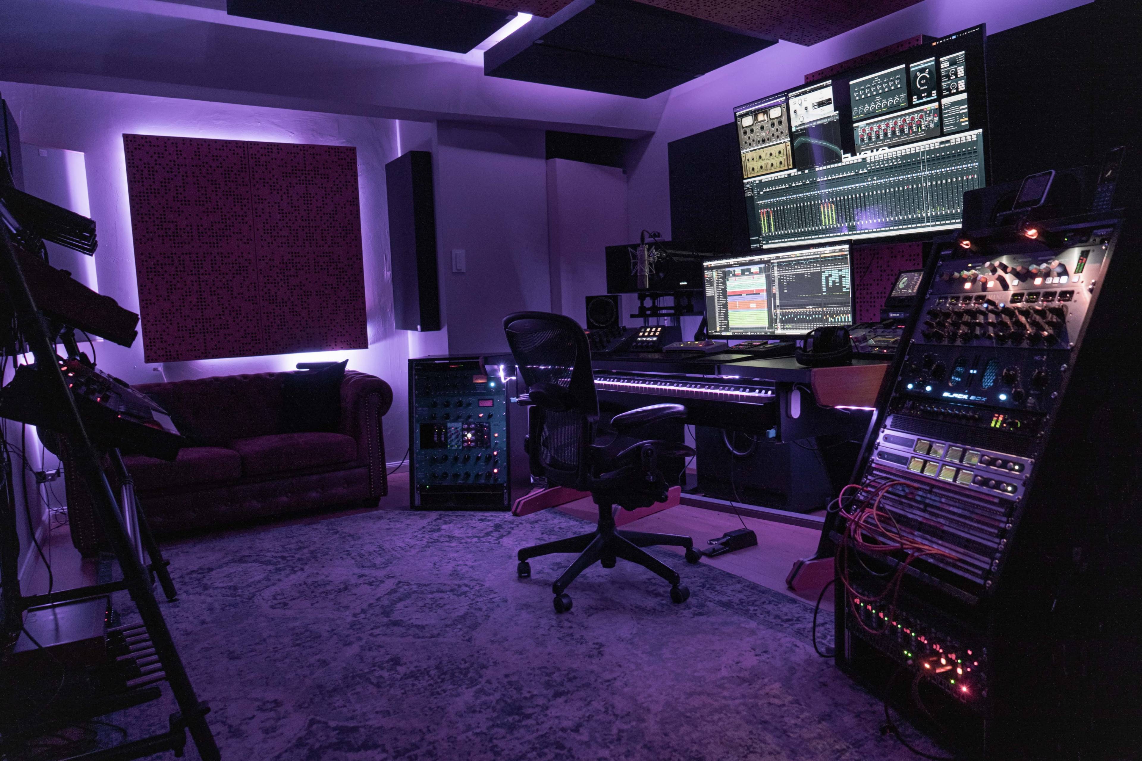 The image shows a modern music studio with a mixing desk, computer monitors, and various audio equipment, illuminated by ambient lighting.