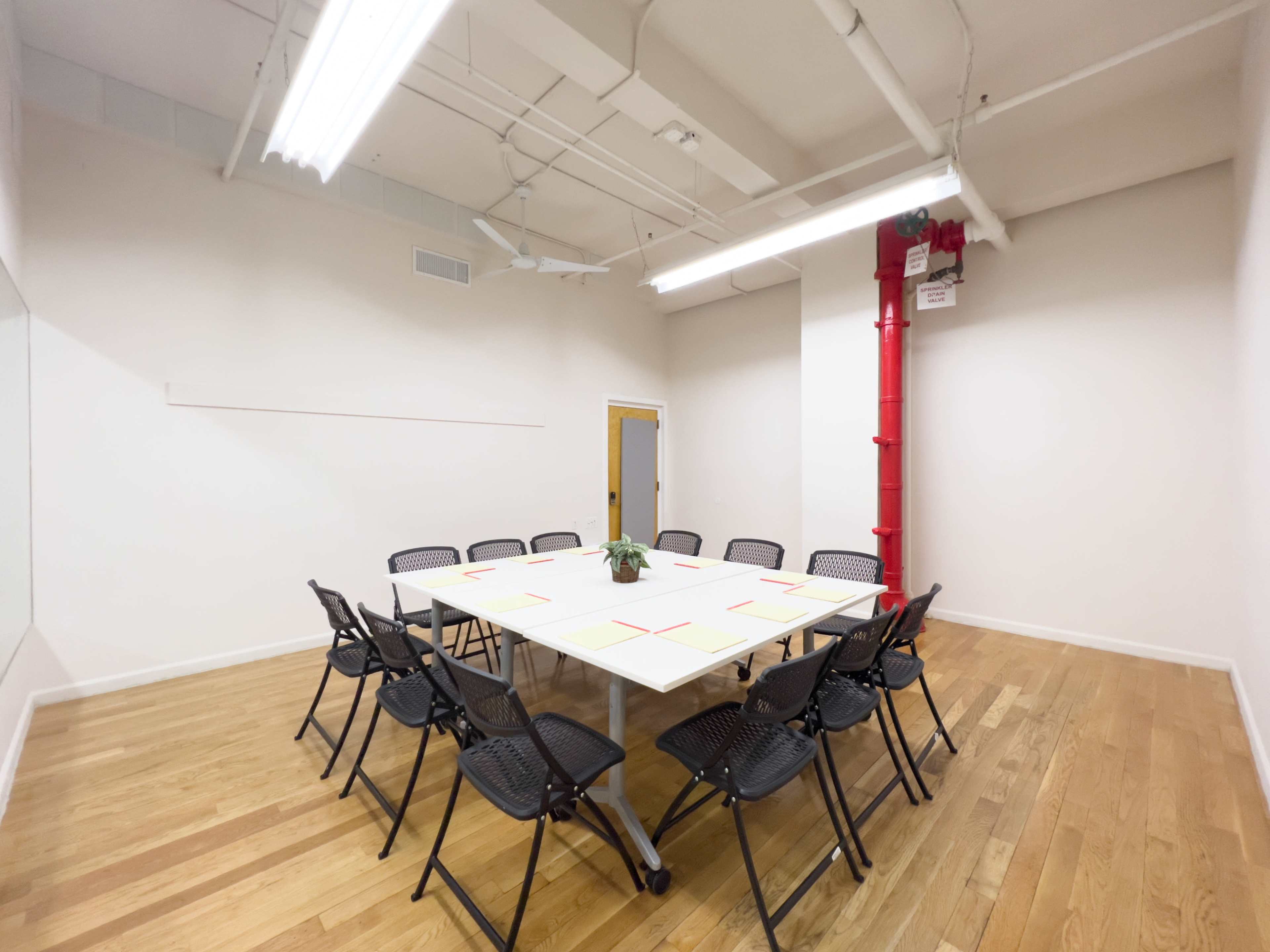 A rectangular table surrounded by black folding chairs sits in a minimalist meeting room with wooden flooring and a red pipe.