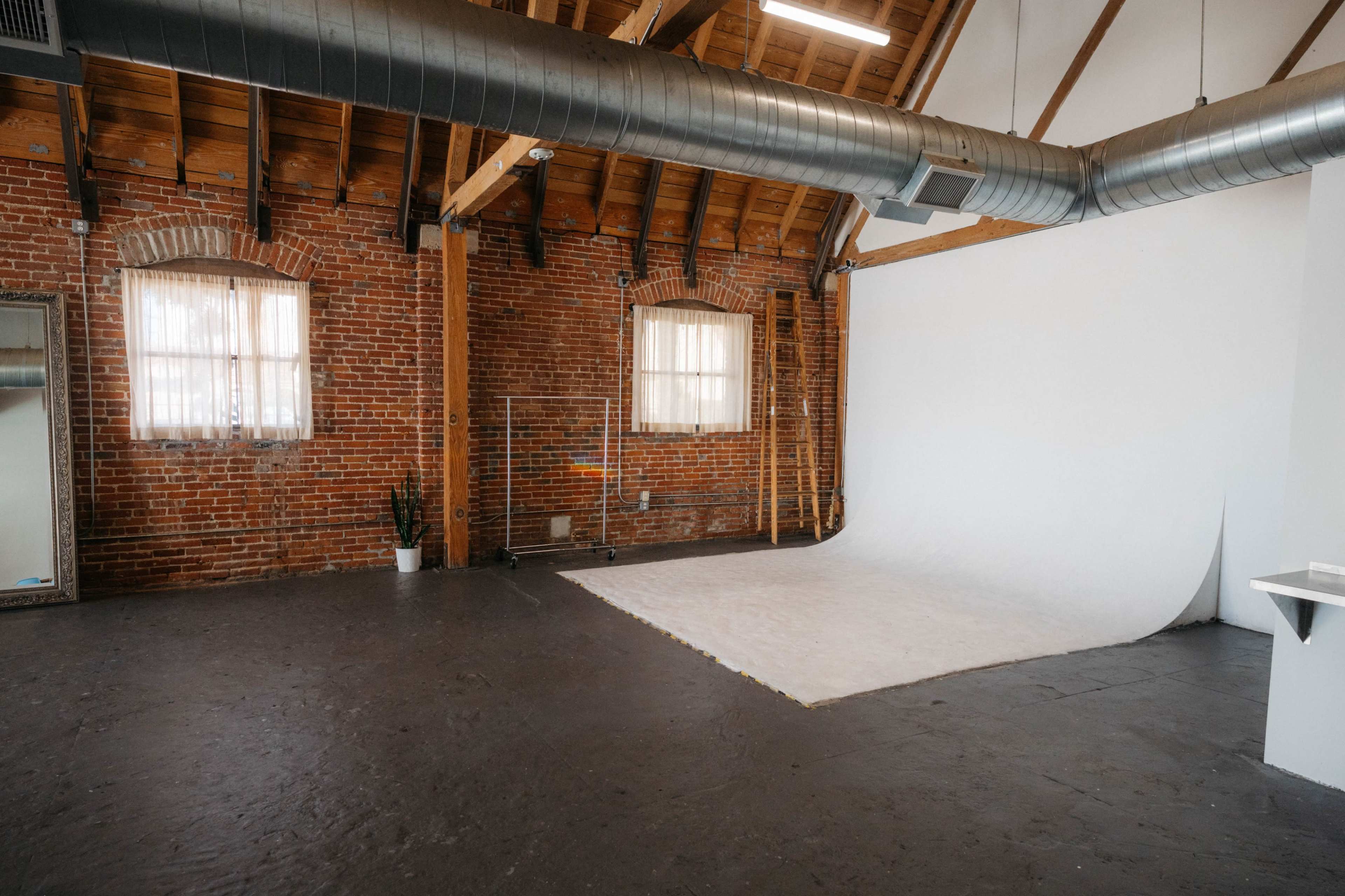 The image shows an empty studio space with exposed brick walls, wooden beams, and a white backdrop set up on the floor.
