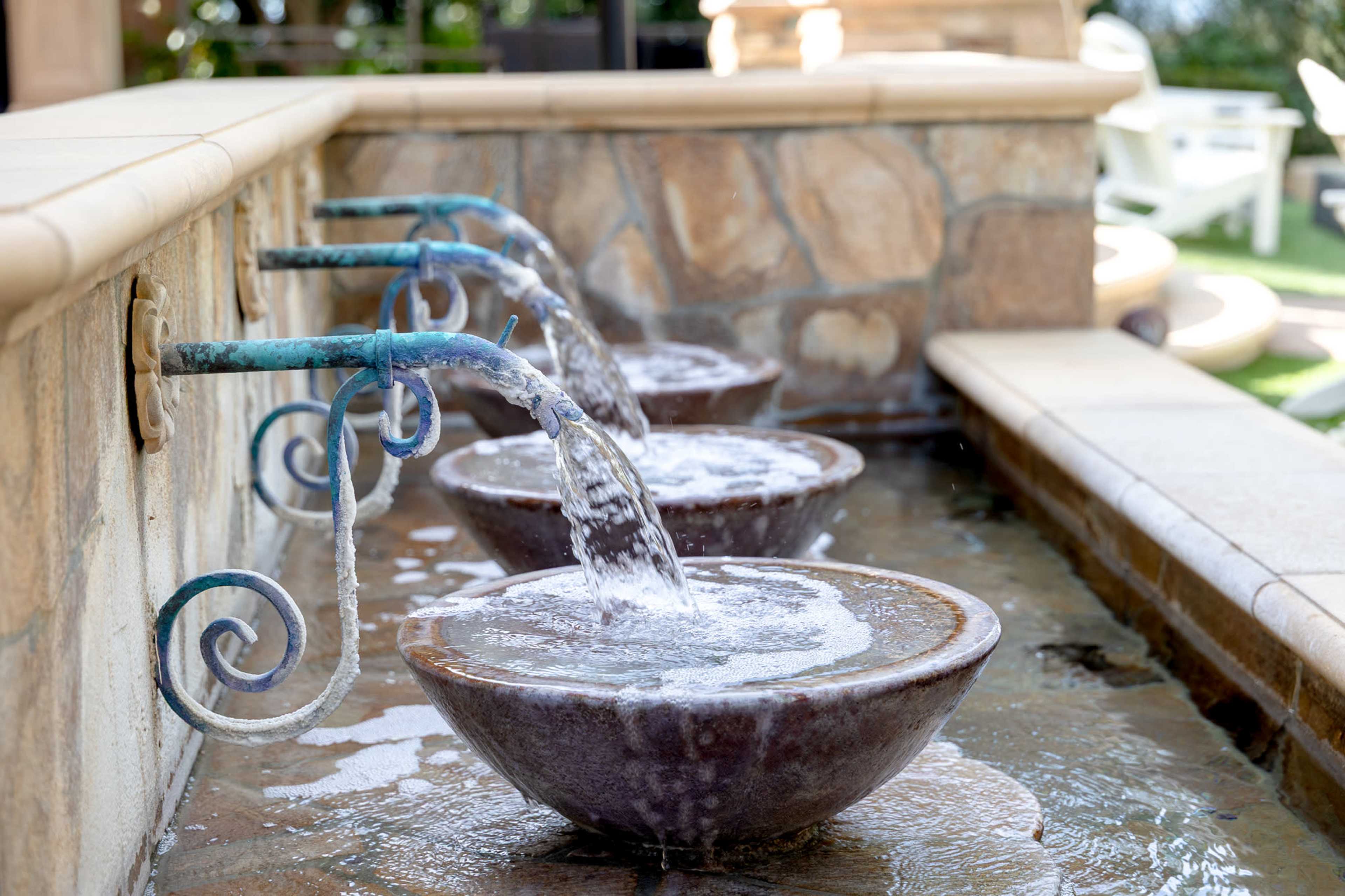 The image shows a series of four ceramic bowls with water flowing into them from decorative spouts, set against a stone wall.