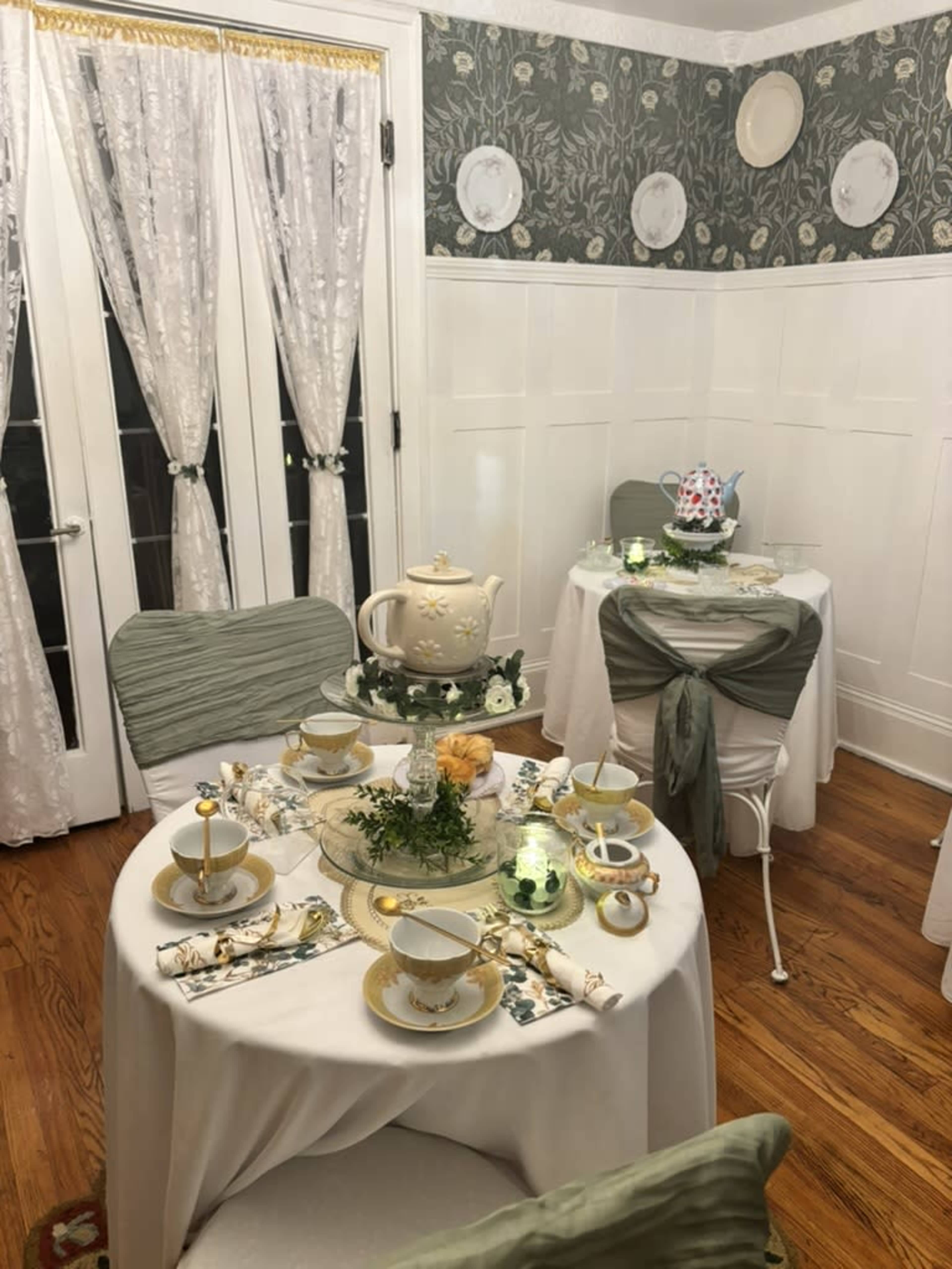 The image shows a cozy dining room setup with two elegantly decorated tables, each featuring fine china, floral centerpieces, and soft green accents.