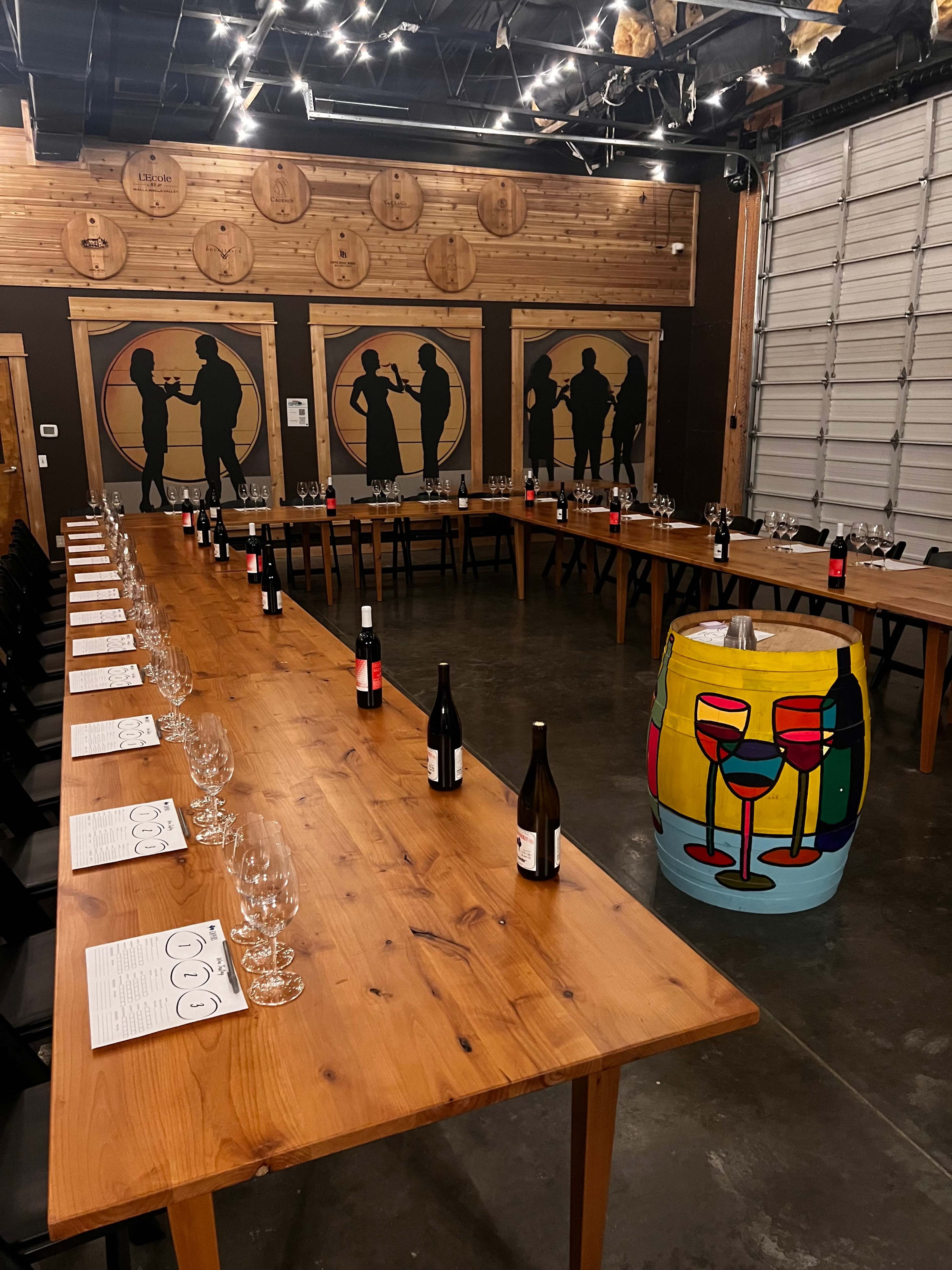 A large wooden dinner table is arranged in a U-shape with wine glasses and bottles, surrounded by silhouetted figures on the walls and a colorful painted barrel at one end.