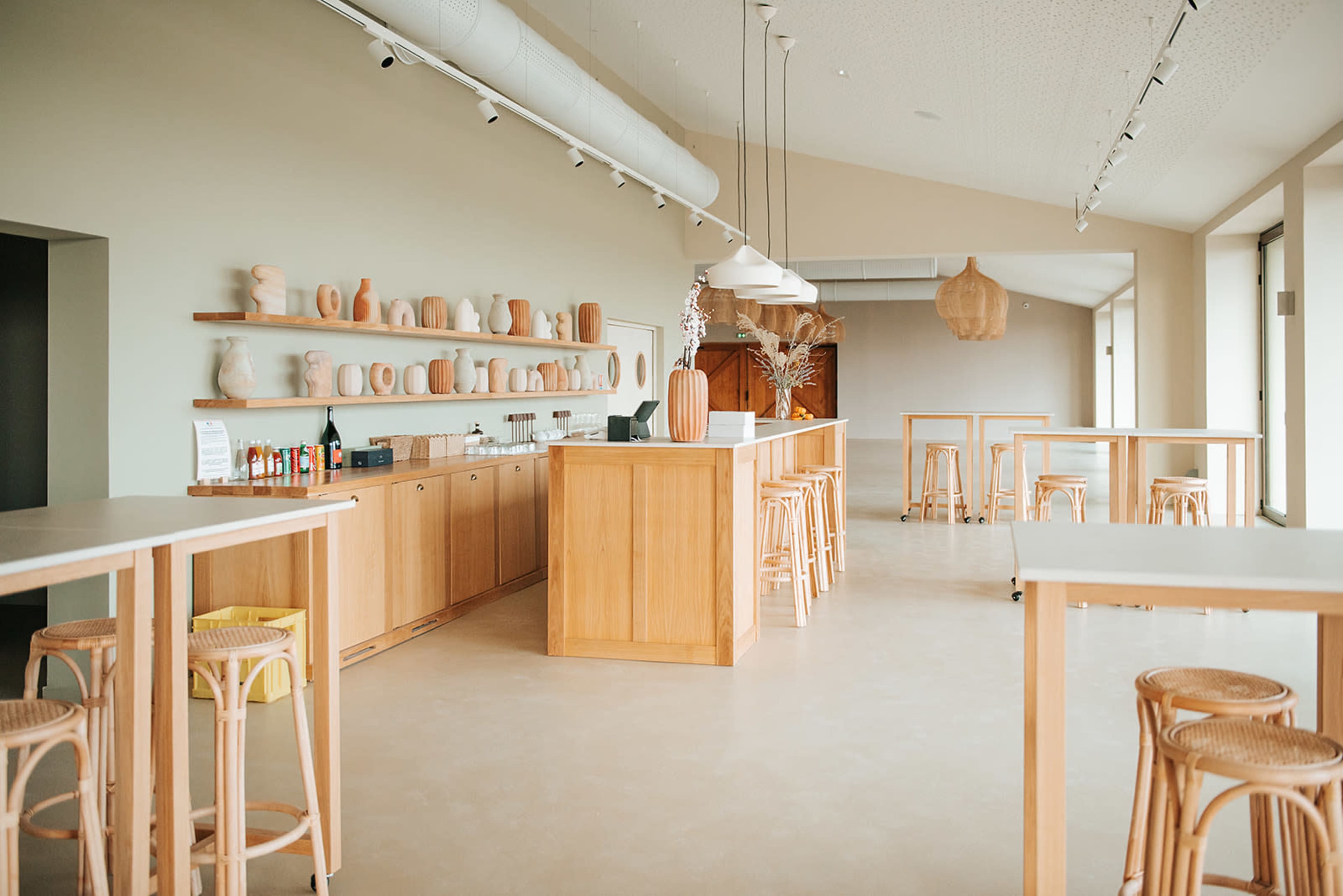 The image depicts a spacious, modern café interior with light-colored walls, wooden furniture, and various decorative vases displayed on a shelf.