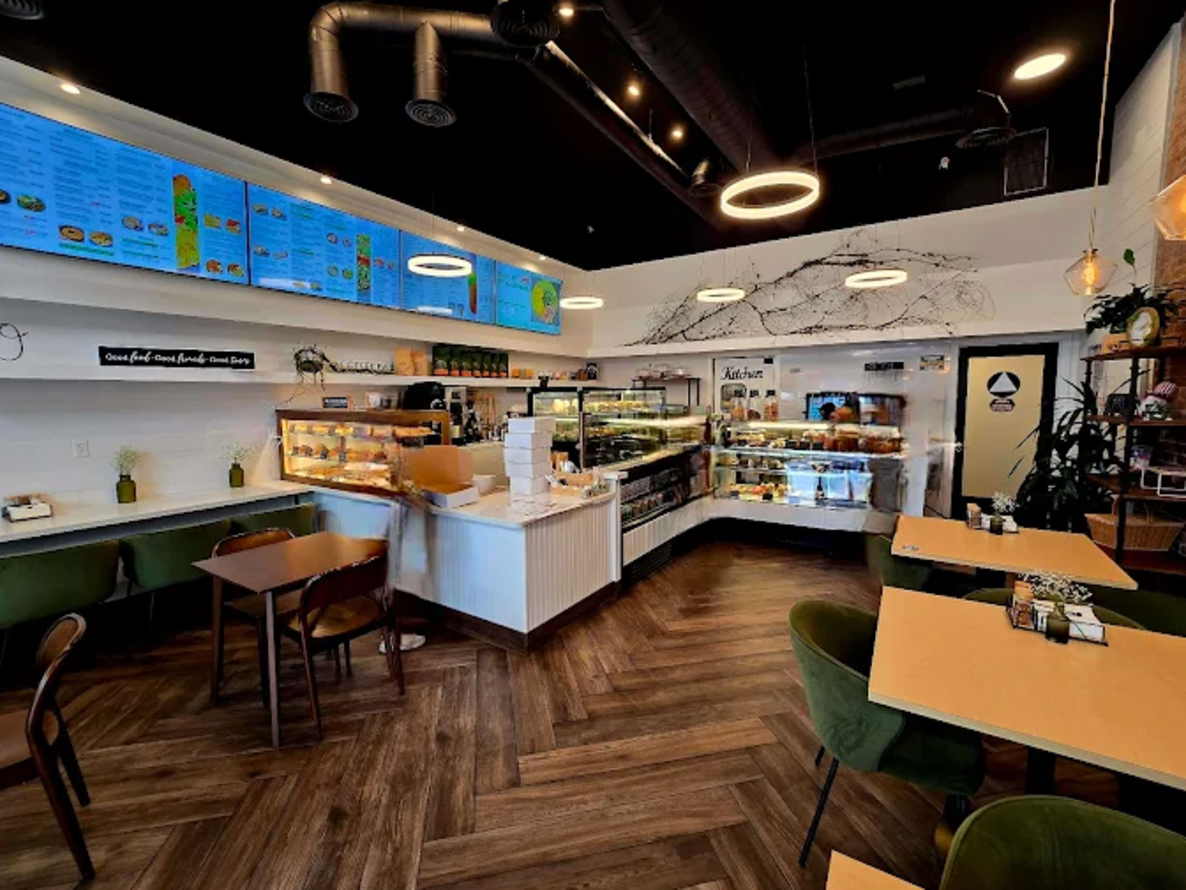 The image shows a modern café interior featuring a bright layout with food display counters, tables for dining, and digital menu boards hanging on the walls.