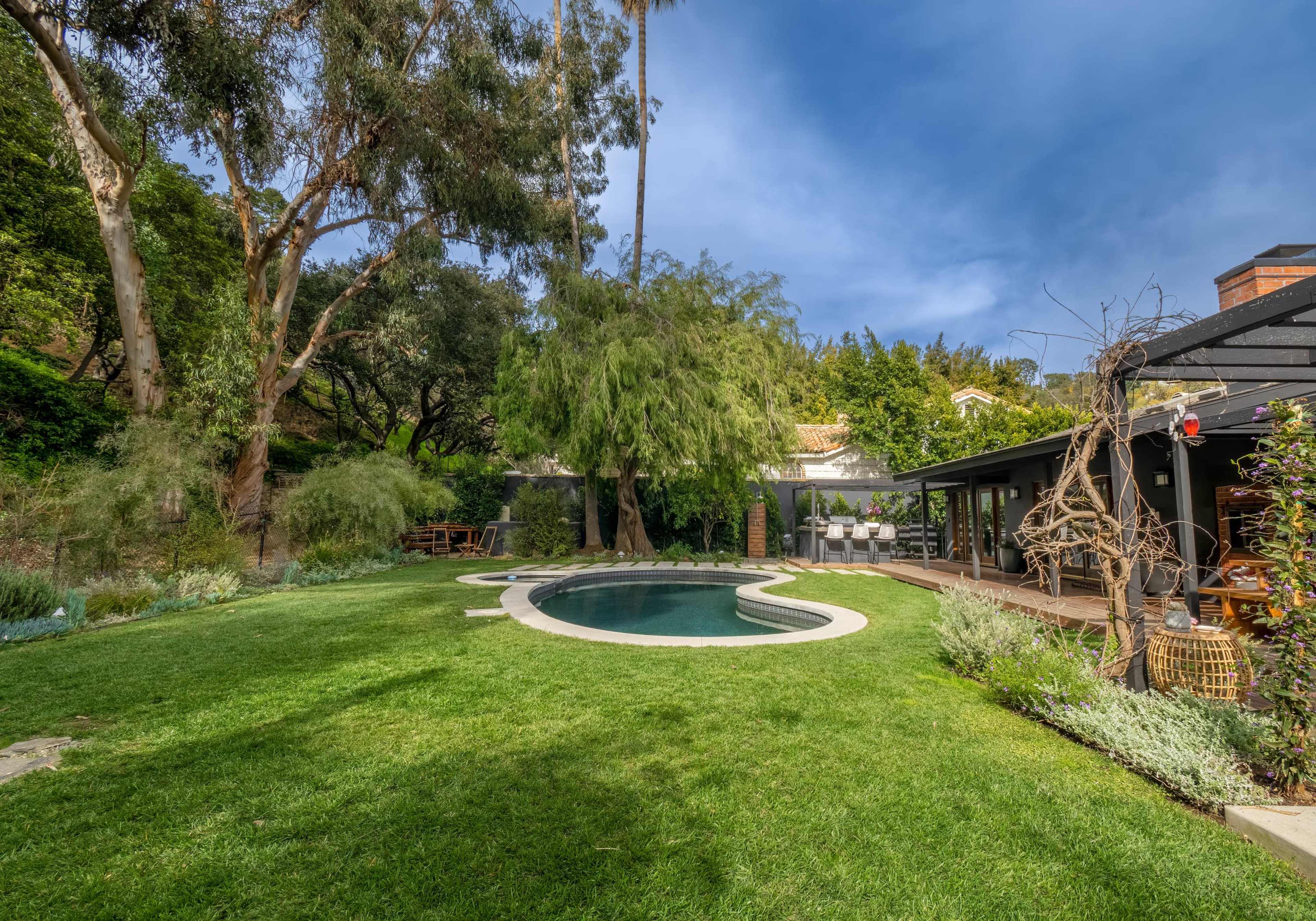 A landscaped backyard features a swimming pool surrounded by grass and trees, with a wooden deck and seating area nearby.