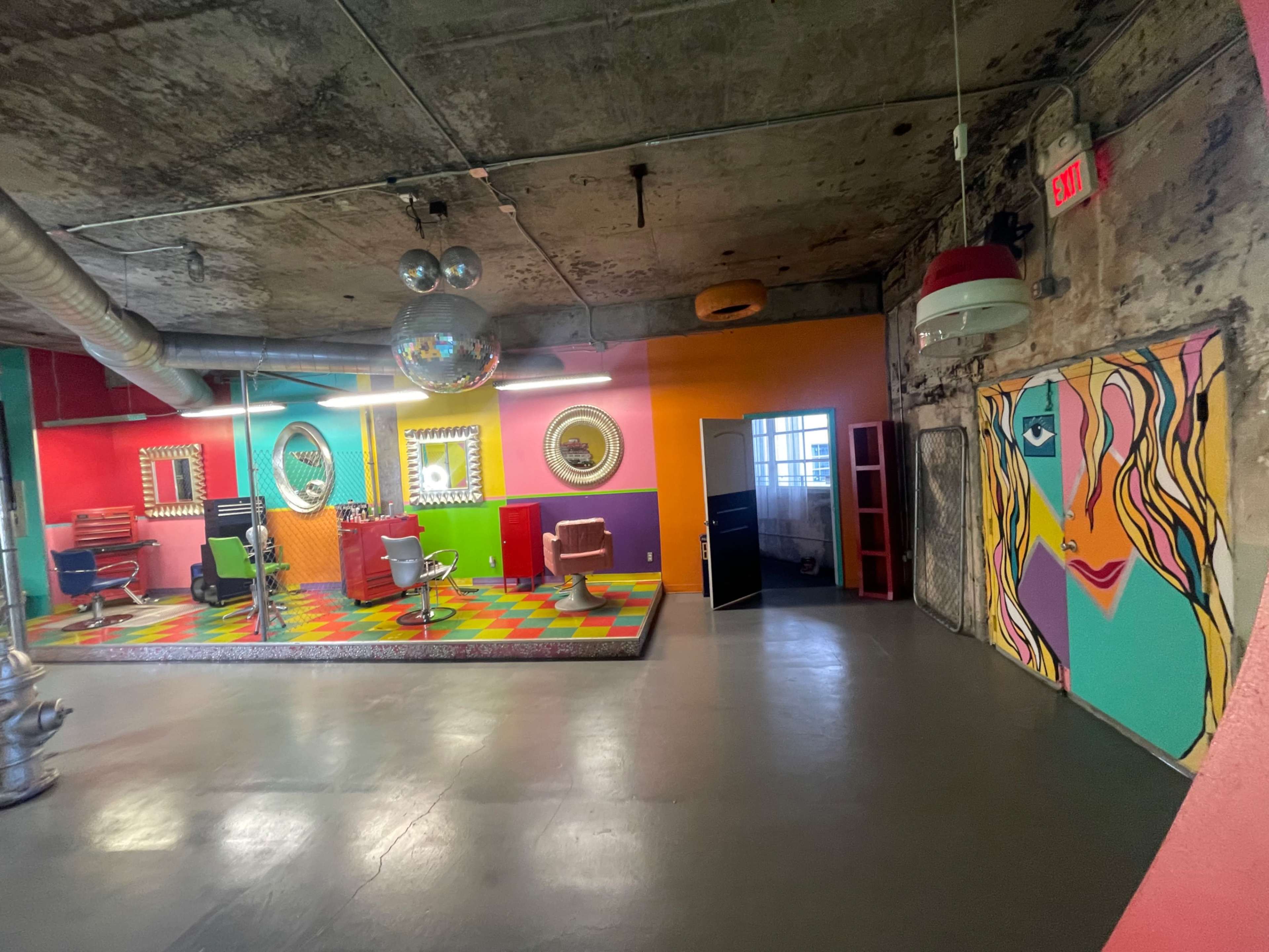A colorful hair salon with a vibrant mural, disco ball, and various styling chairs against a backdrop of textured concrete walls.
