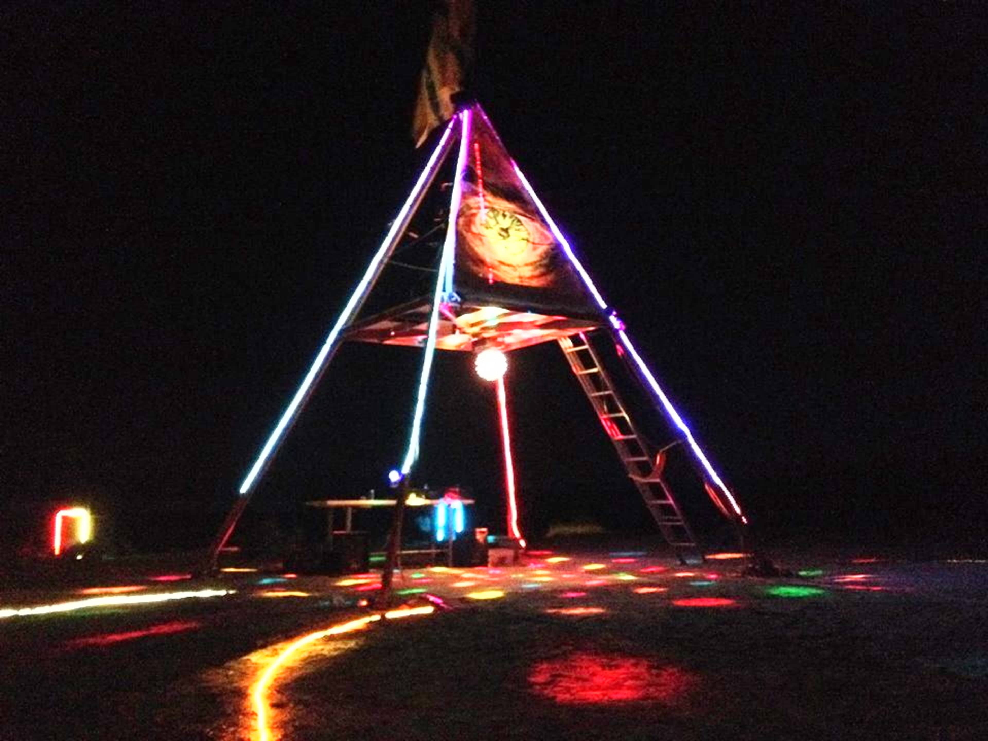 A brightly lit pyramid structure with colorful lights illuminates the ground in a dark outdoor setting.