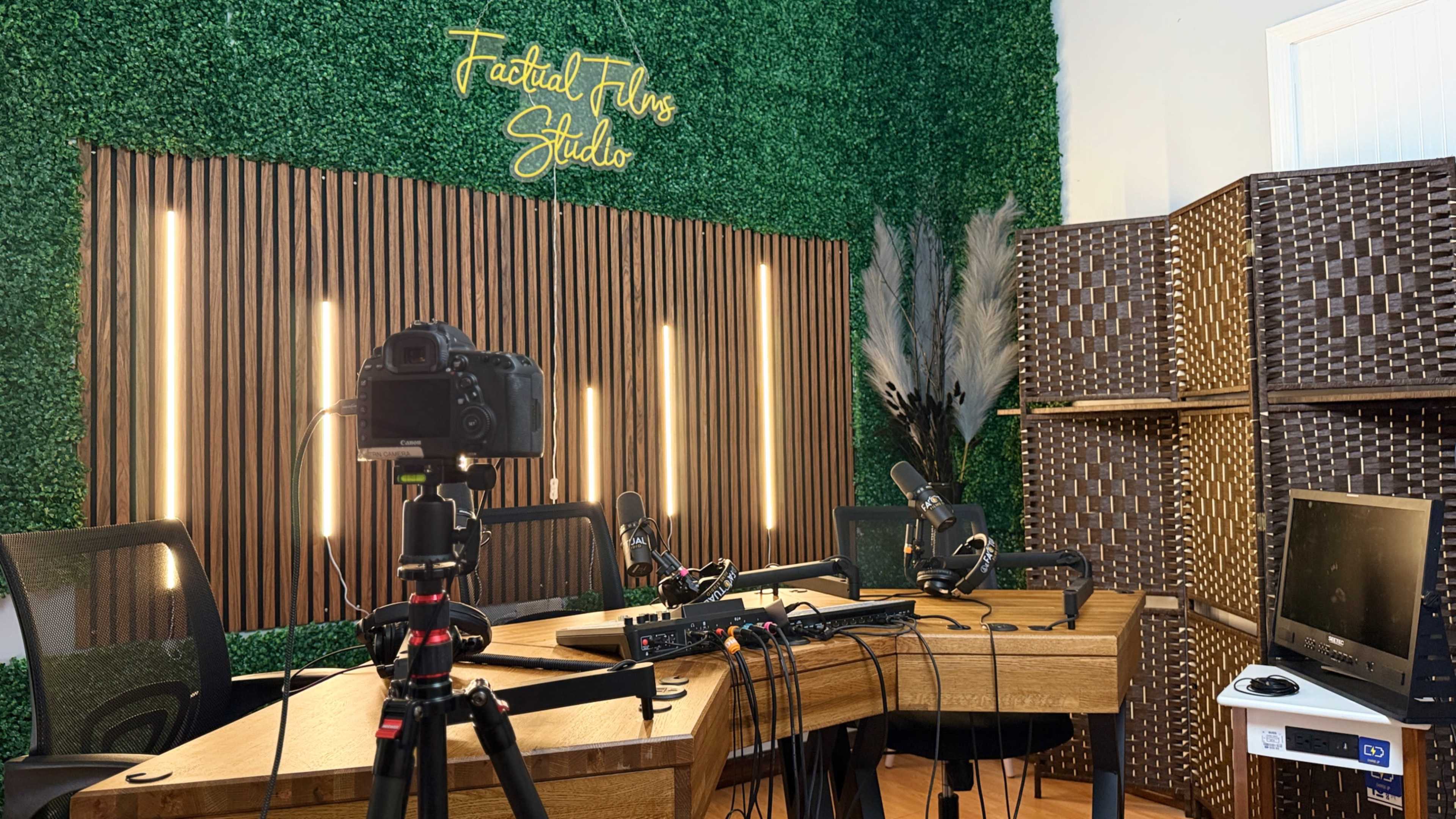 The image shows a podcast studio featuring a wooden table with microphones, a camera on a tripod, and a backdrop of artificial greenery and decorative screens.