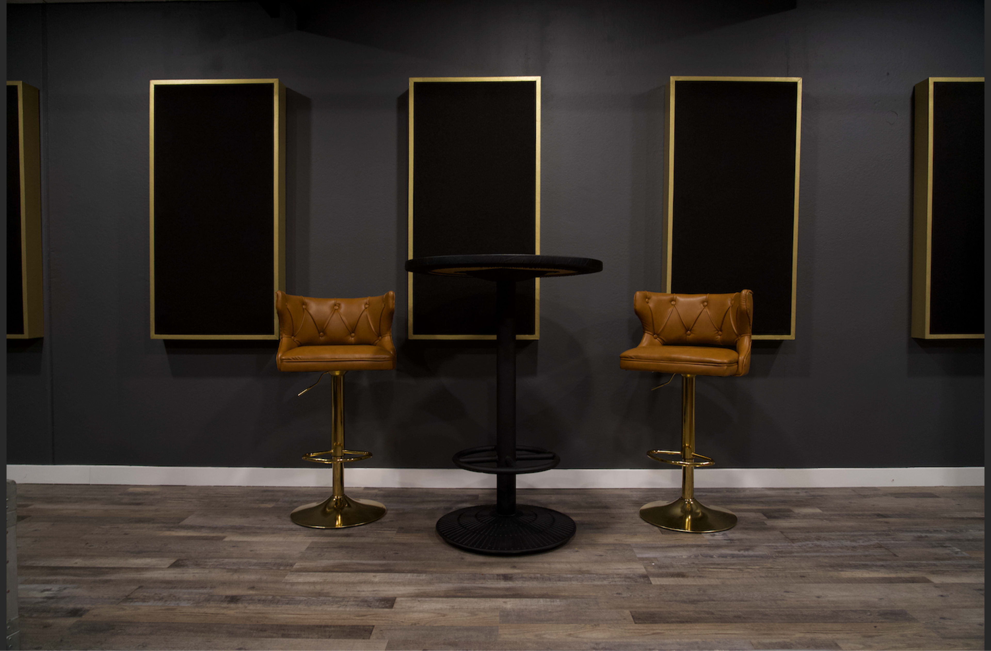 Two golden bar stools and a round black table set against a gray wall with three framed black panels.