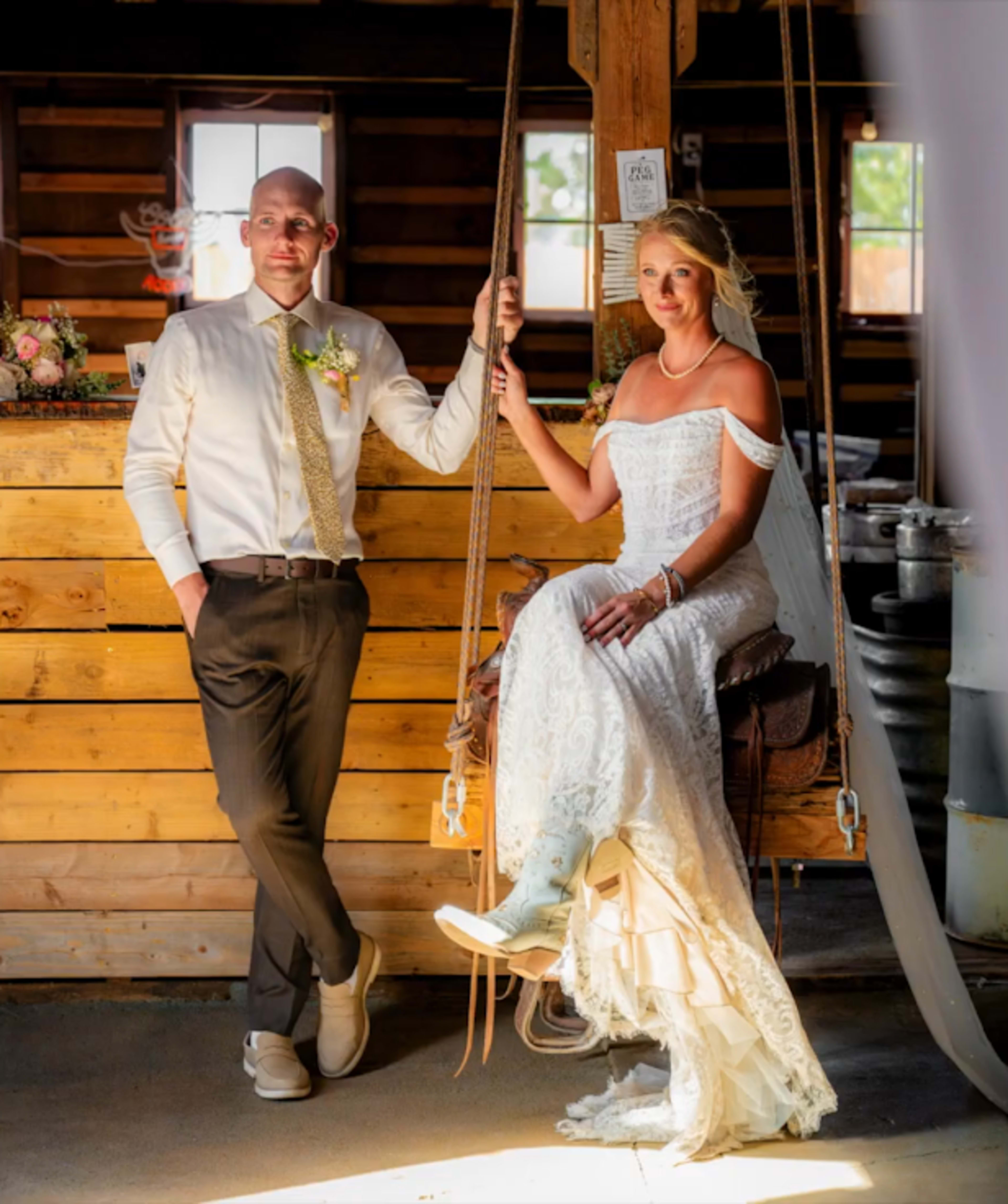 A bride in a white lace gown sits on a swing while a groom in a white shirt and tie stands beside her in a rustic wooden interior.