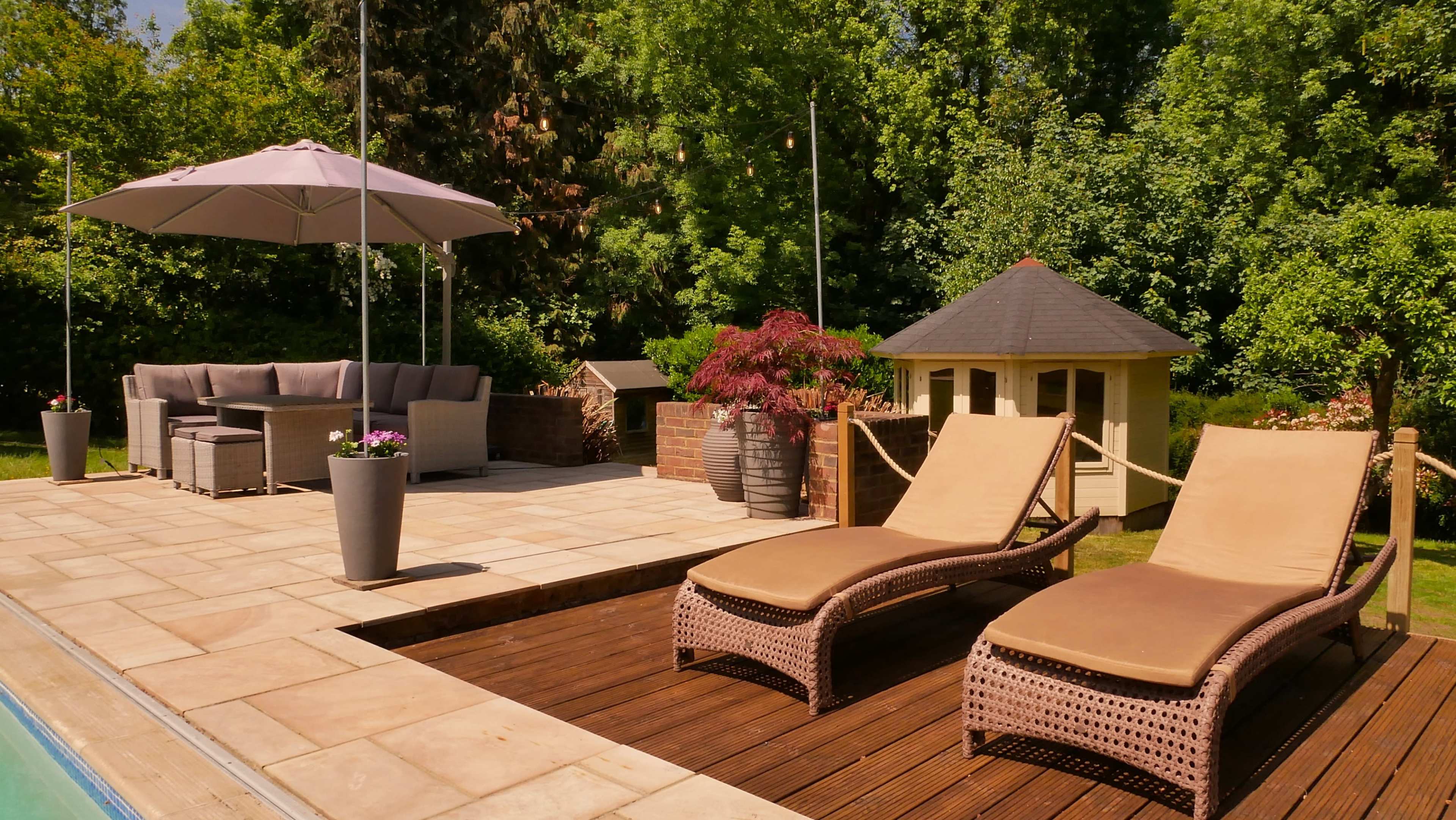 The scene features a patio with two lounge chairs beside a swimming pool, a shaded seating area, and a small garden house surrounded by greenery.