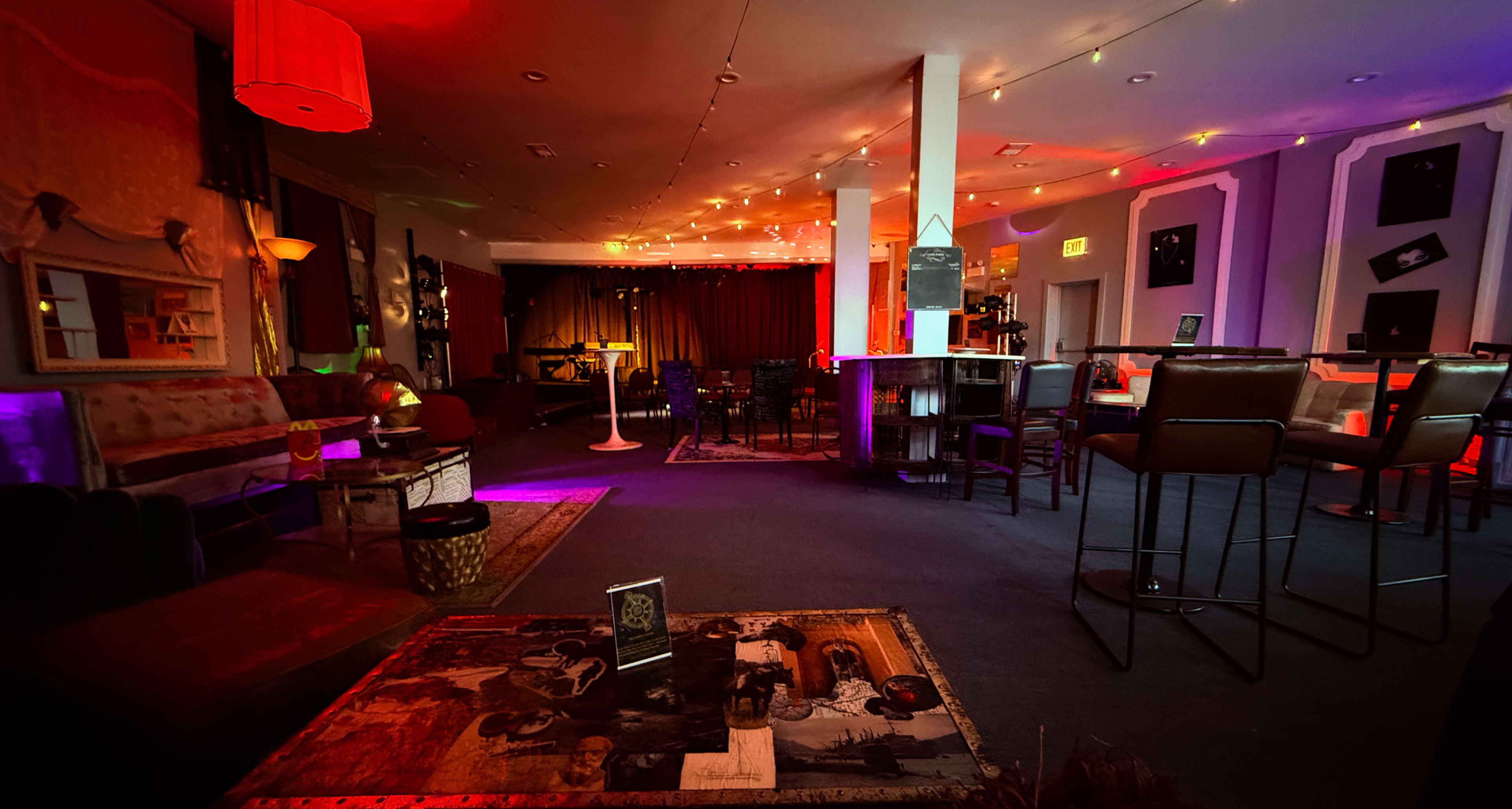 The image shows a dimly lit lounge or bar area adorned with colorful lighting, featuring various seating arrangements and a stage in the background.