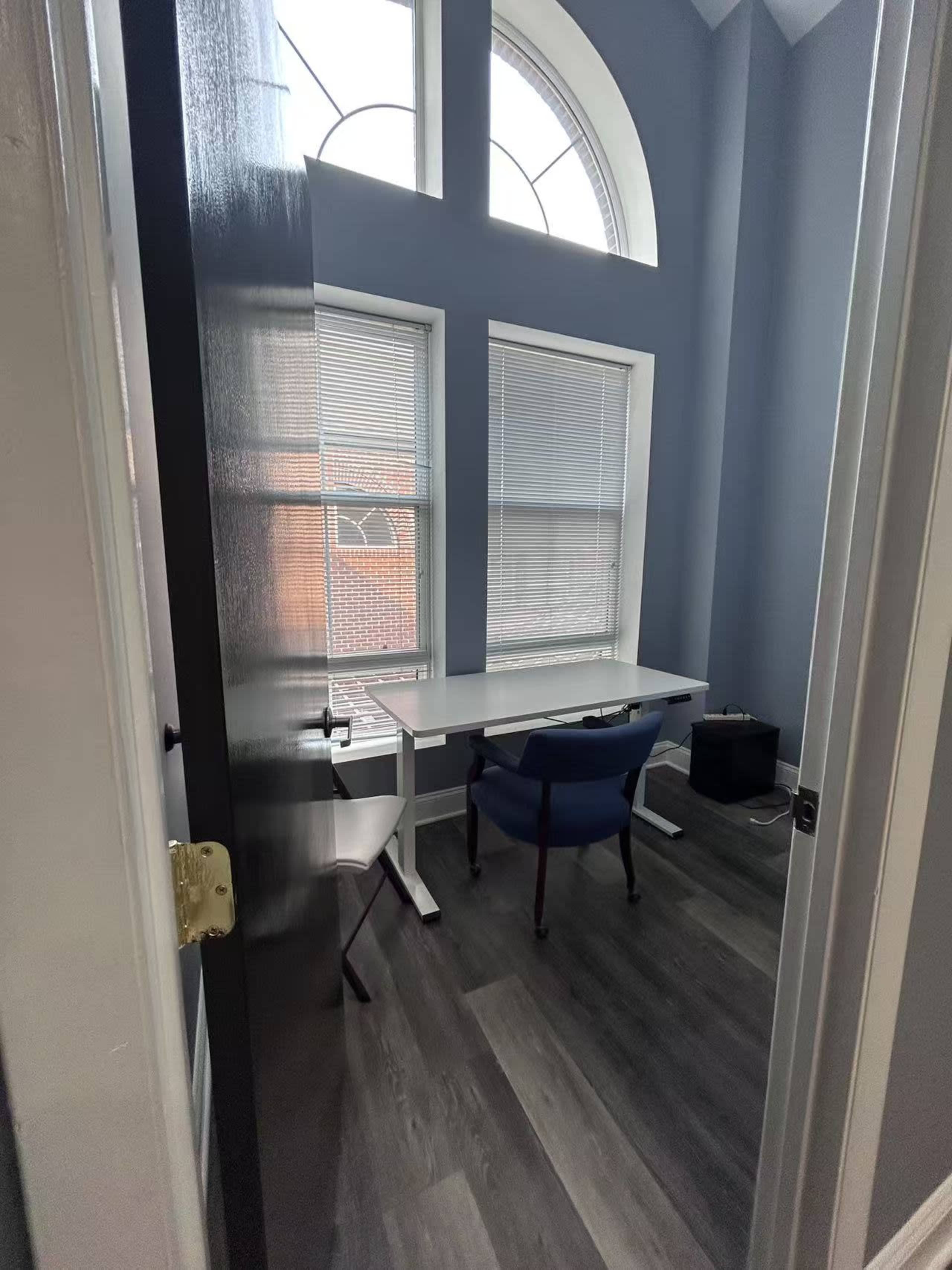The image shows a small office space featuring a white desk and a blue chair, with large windows allowing natural light to enter.