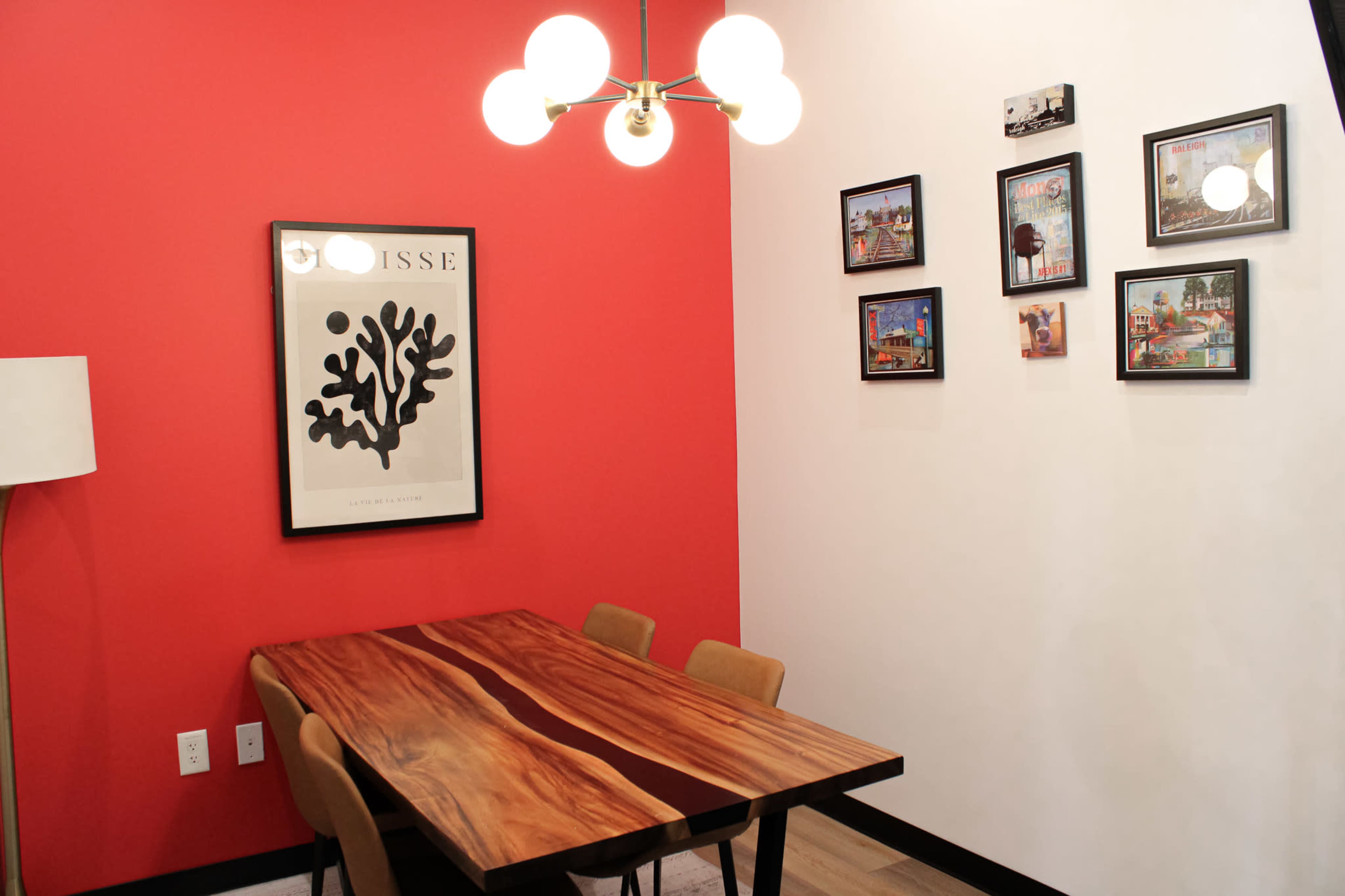 The image shows a dining area with a wooden table, six chairs, a red accent wall featuring a framed artwork, and several framed pictures on the adjacent wall.