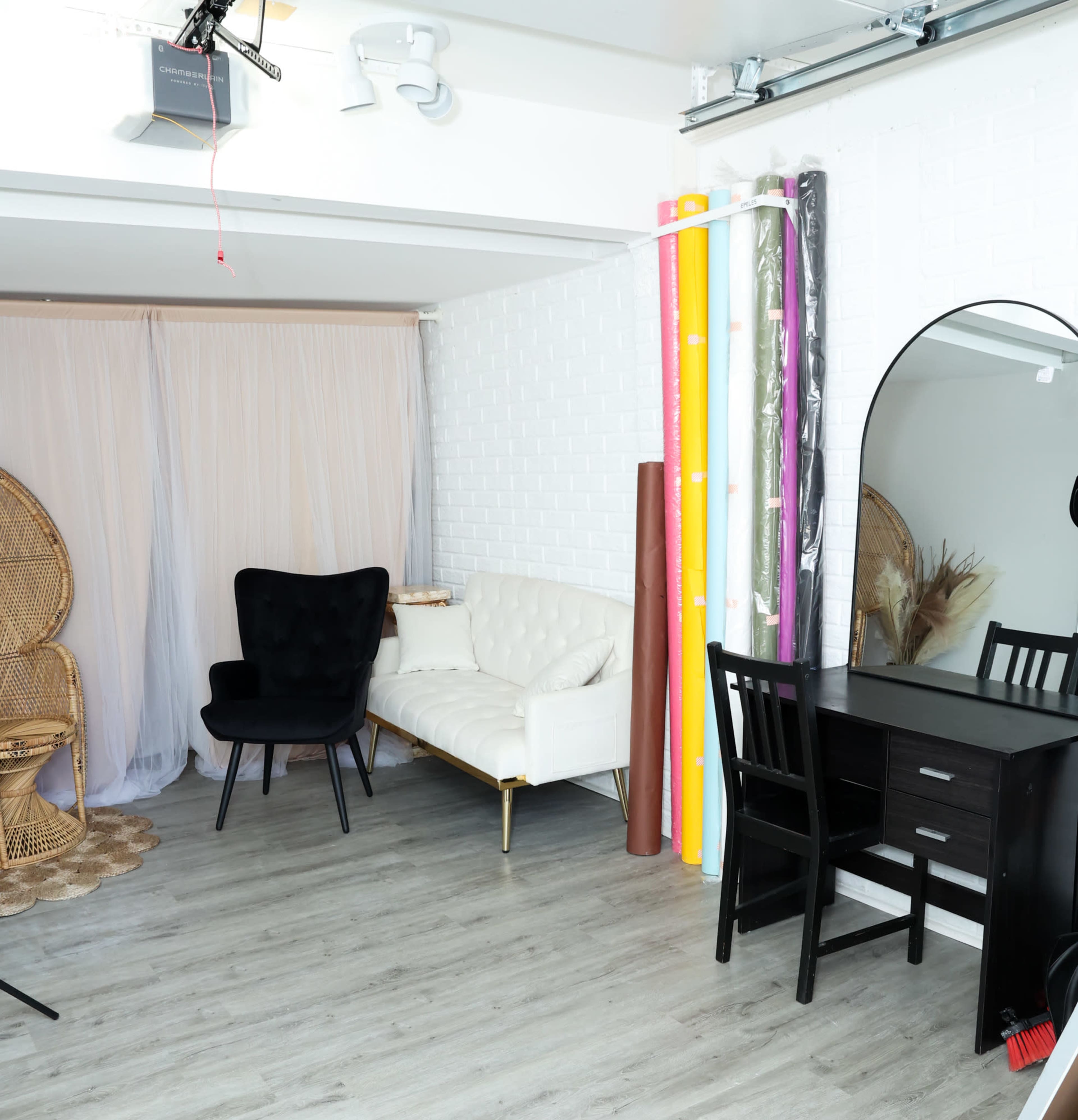 A spacious room featuring a white sofa, two chairs, a dressing mirror, and colorful backdrop rolls against a white brick wall.