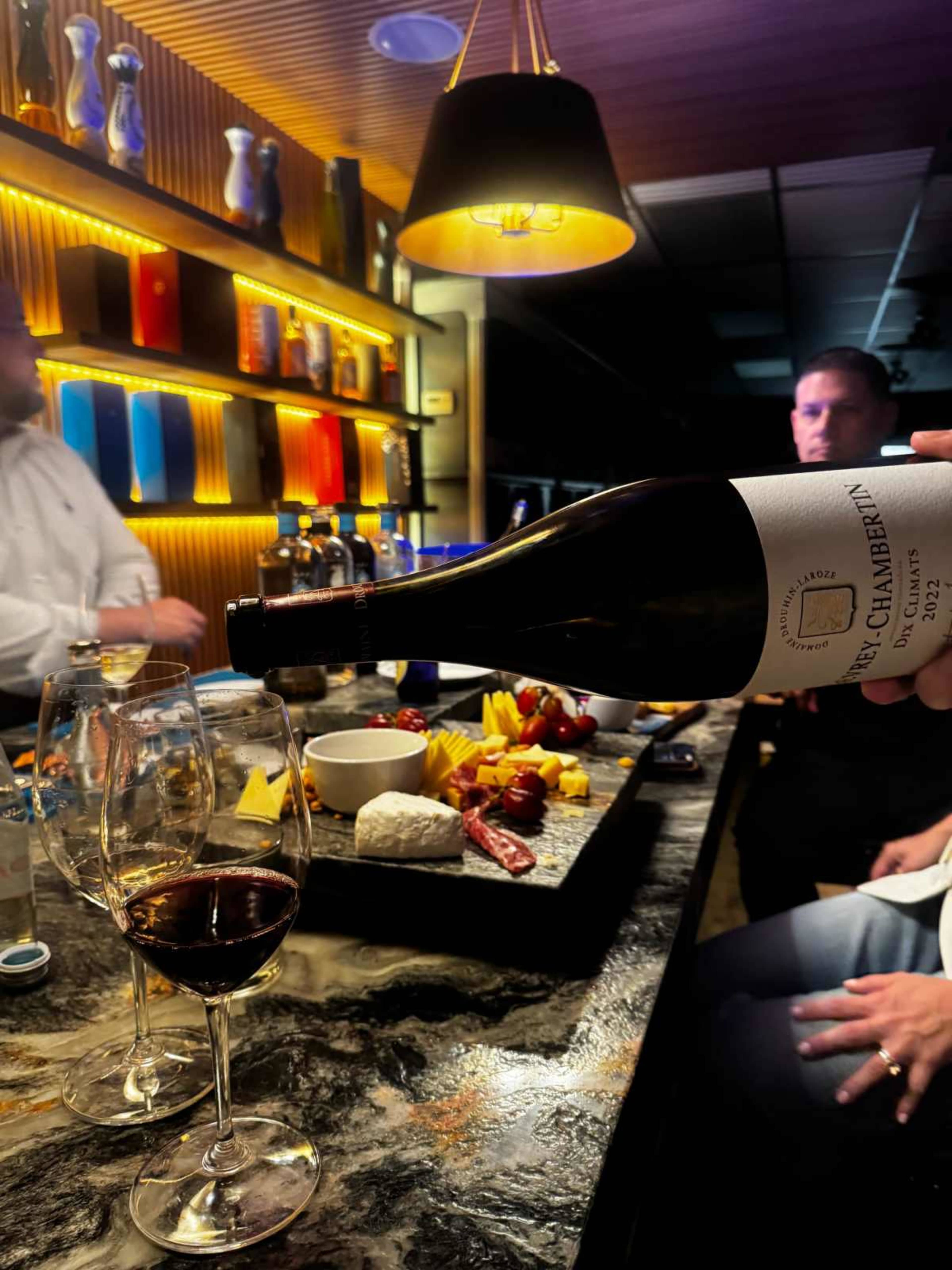 A hand pours red wine from a bottle into glasses on a bar counter adorned with a platter of cheeses, meats, and fruits, with blurred figures in the background.