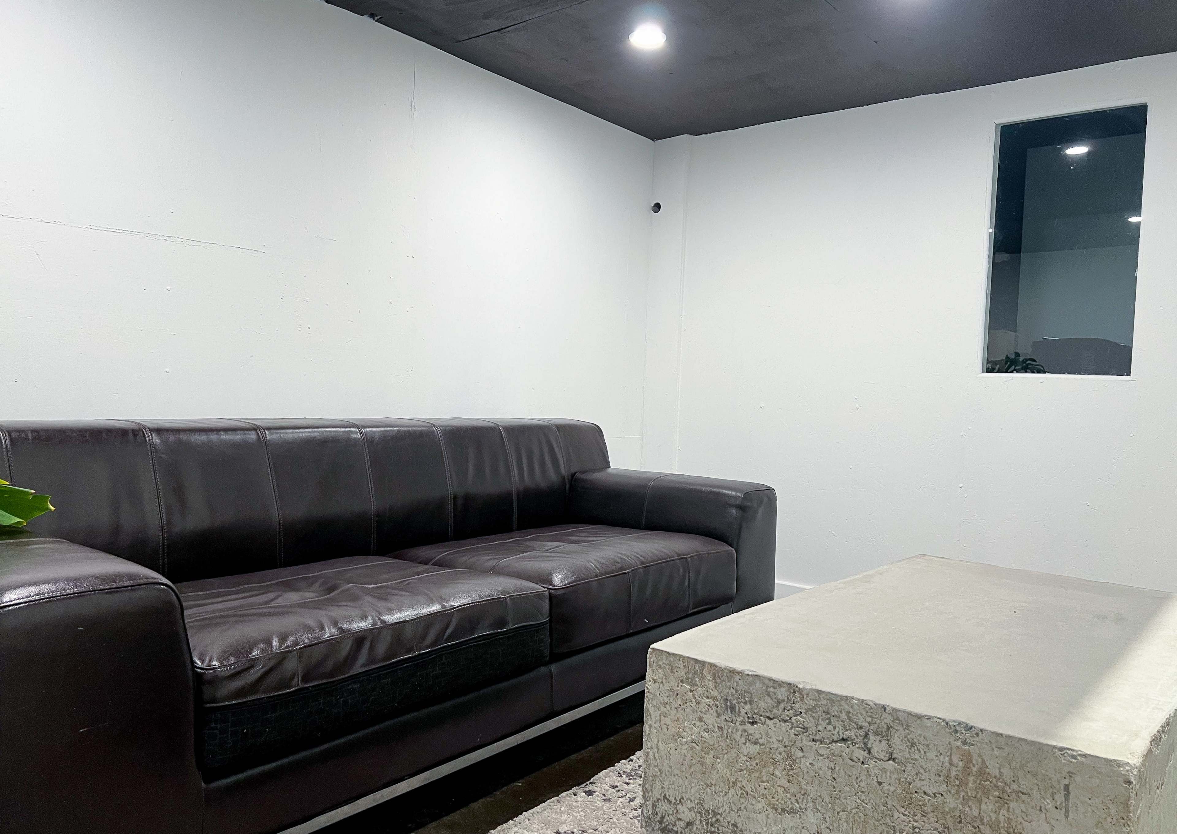 A brown leather sofa facing a concrete coffee table in a minimalist room with a white wall and a small window.