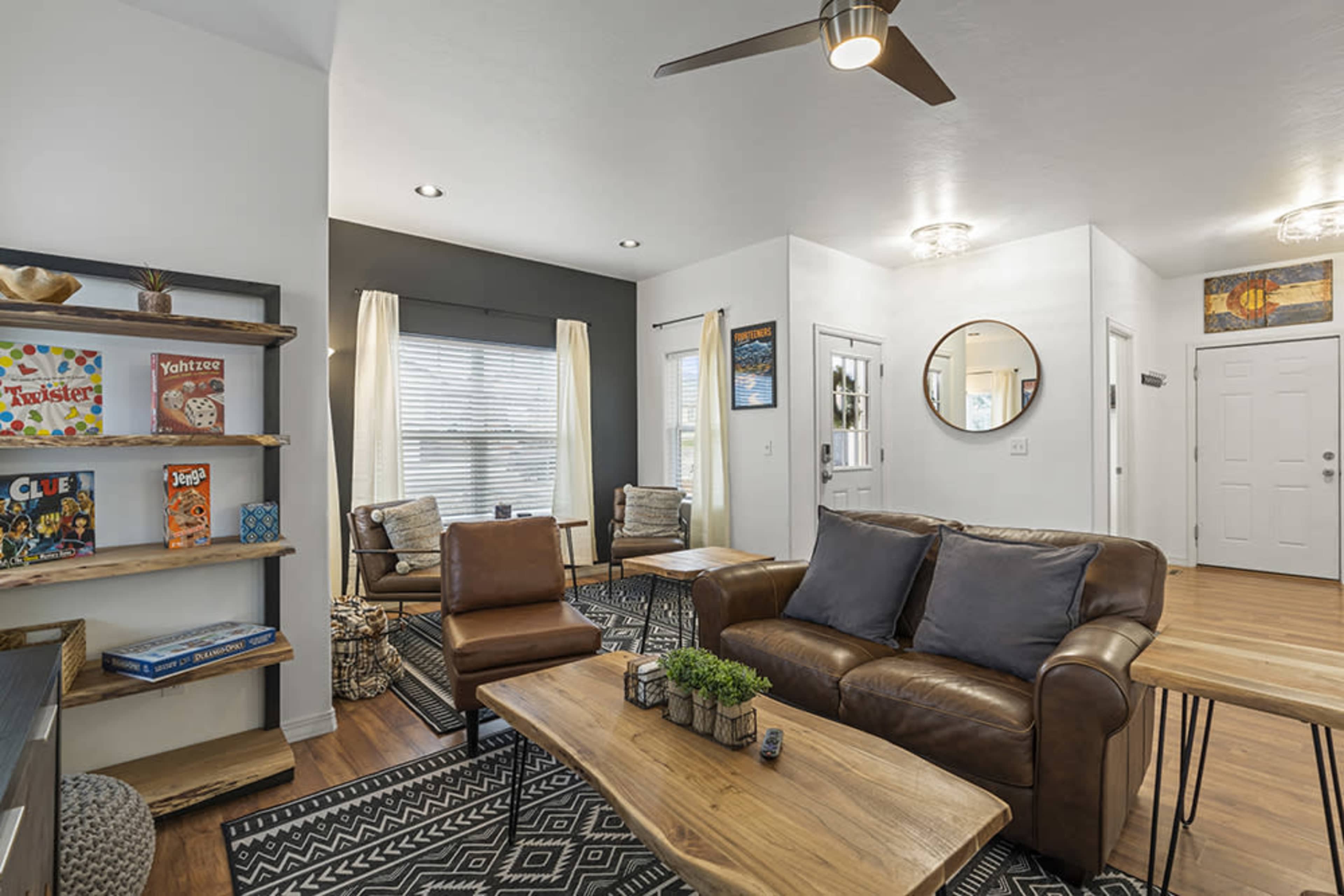 The living room features a leather sofa, wooden coffee table, and a bookshelf displaying board games, all set against a backdrop of large windows with natural light.