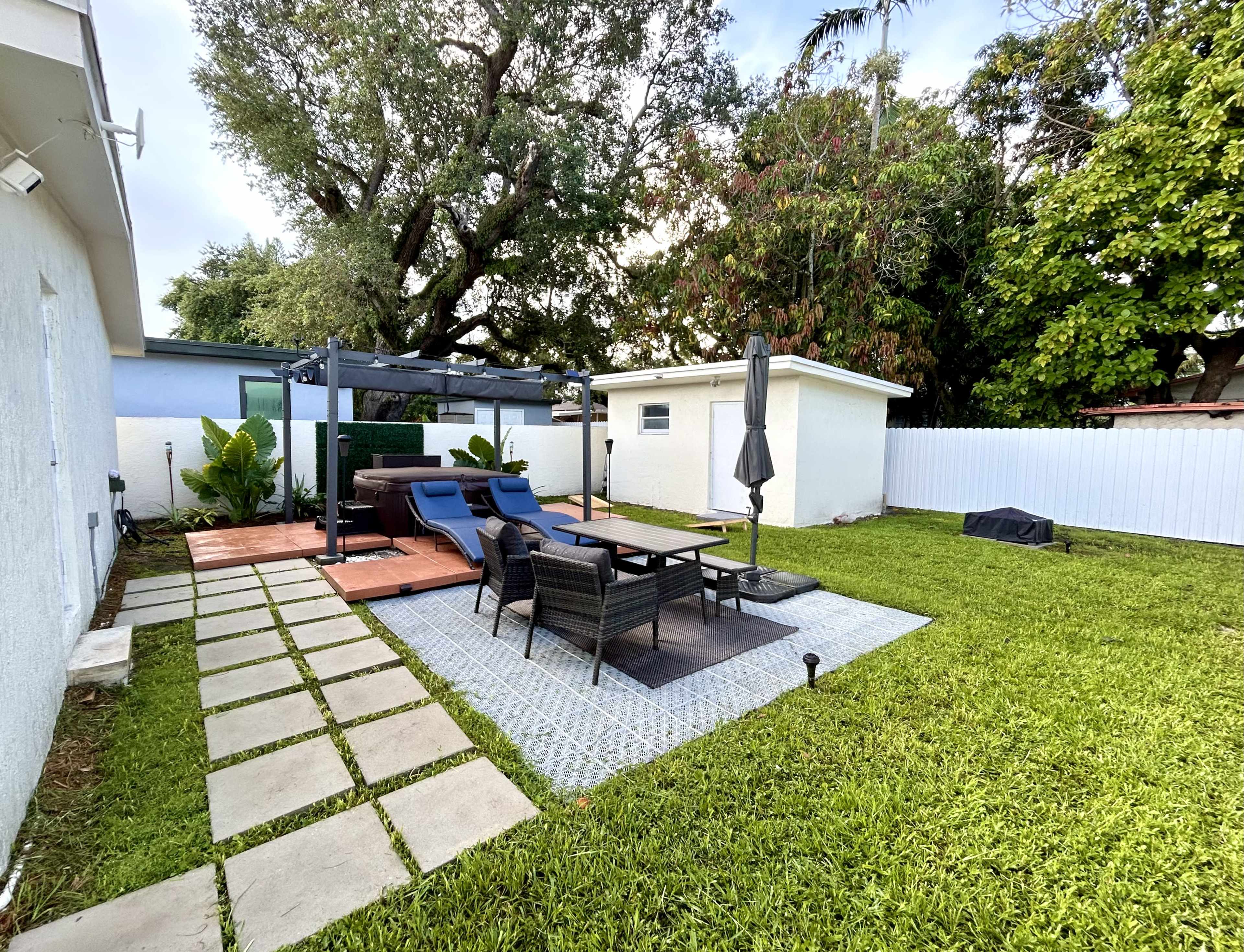 The image shows a backyard featuring a patio with lounge chairs, a table, and a small shed surrounded by greenery and a white fence.