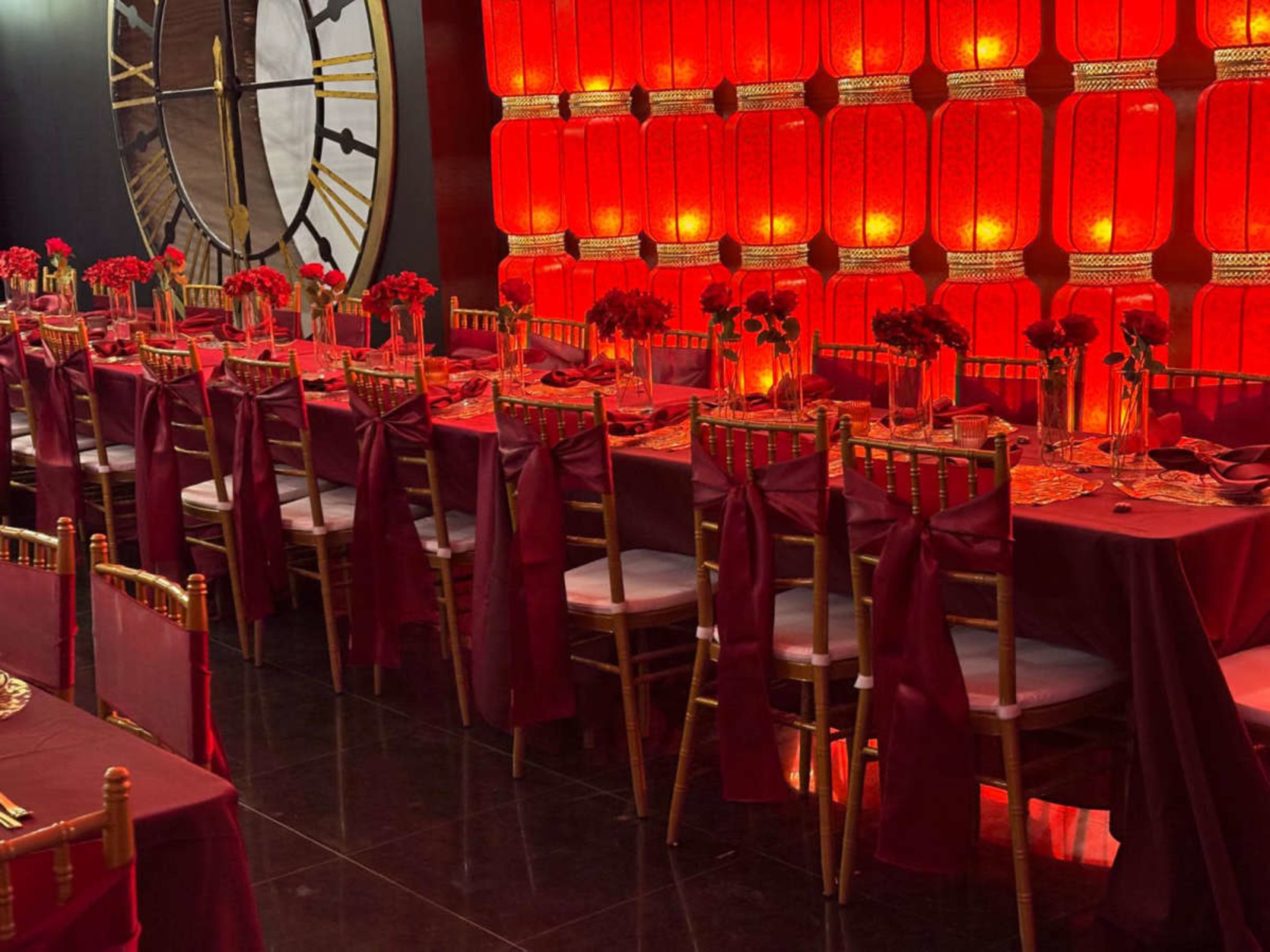 The image shows a banquet hall featuring long tables covered in deep red tablecloths, adorned with floral centerpieces and surrounded by gold chairs, set against a backdrop of red glowing lanterns.