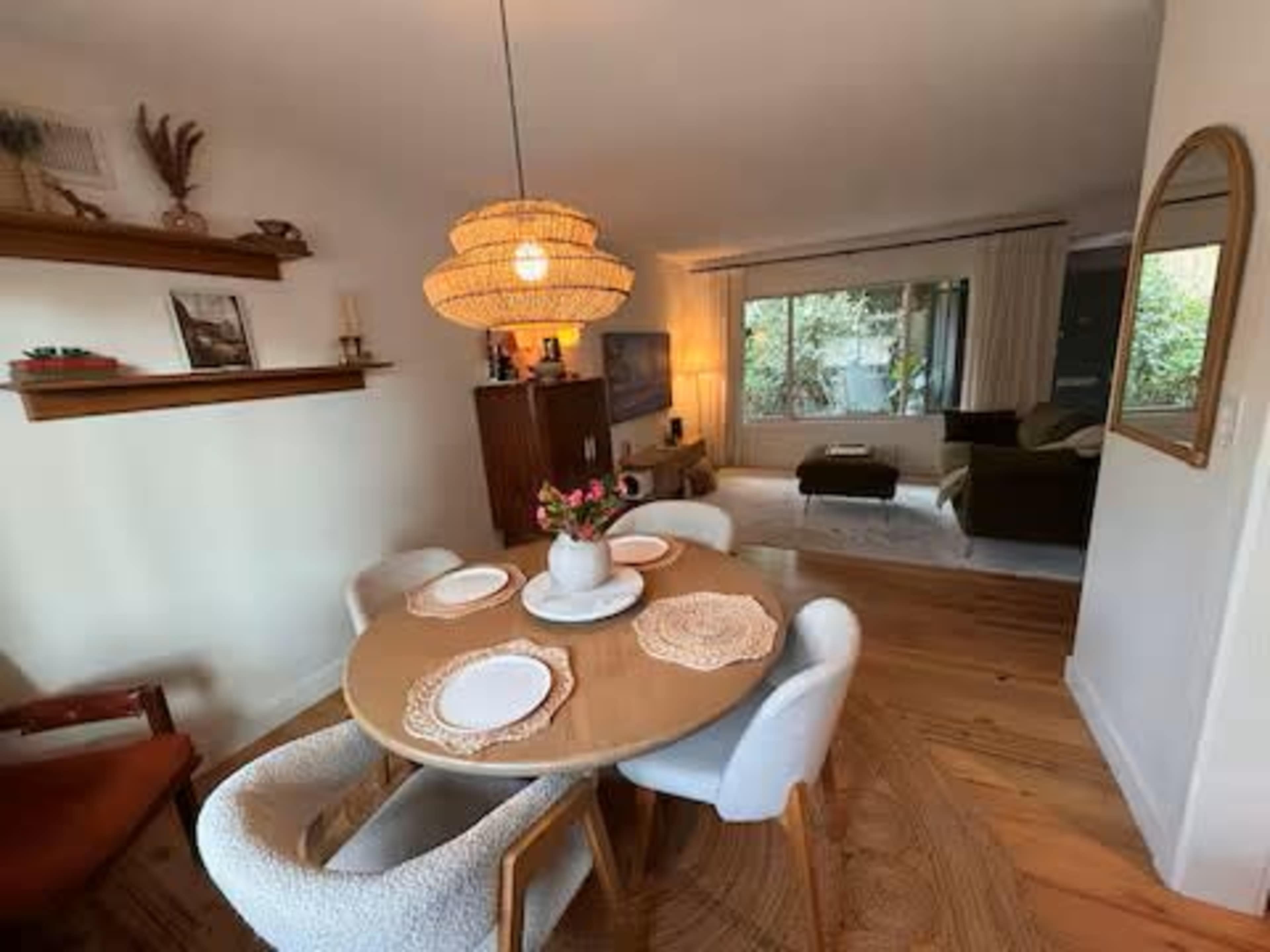 A dining area with a round wooden table set for four, featuring a large woven pendant light, and a cozy living space visible in the background.