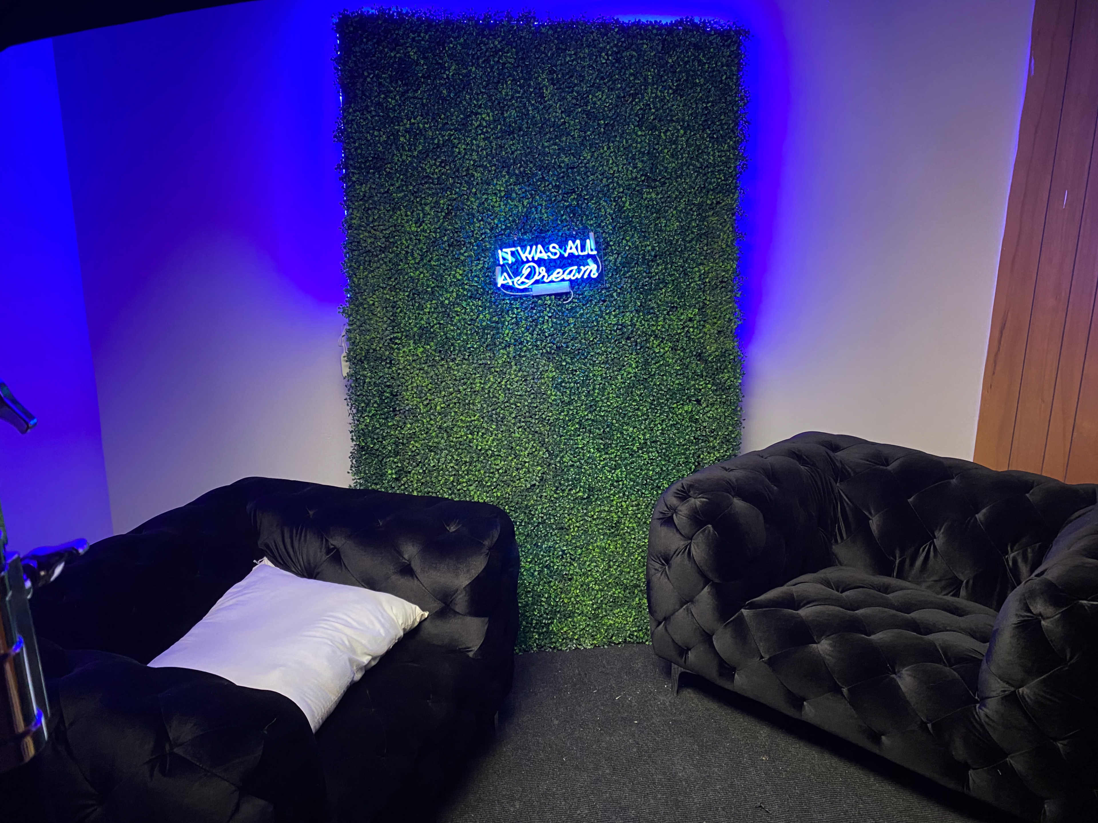A small room features two black tufted sofas facing a green wall adorned with a neon sign that reads, "It Was All a Dream."
