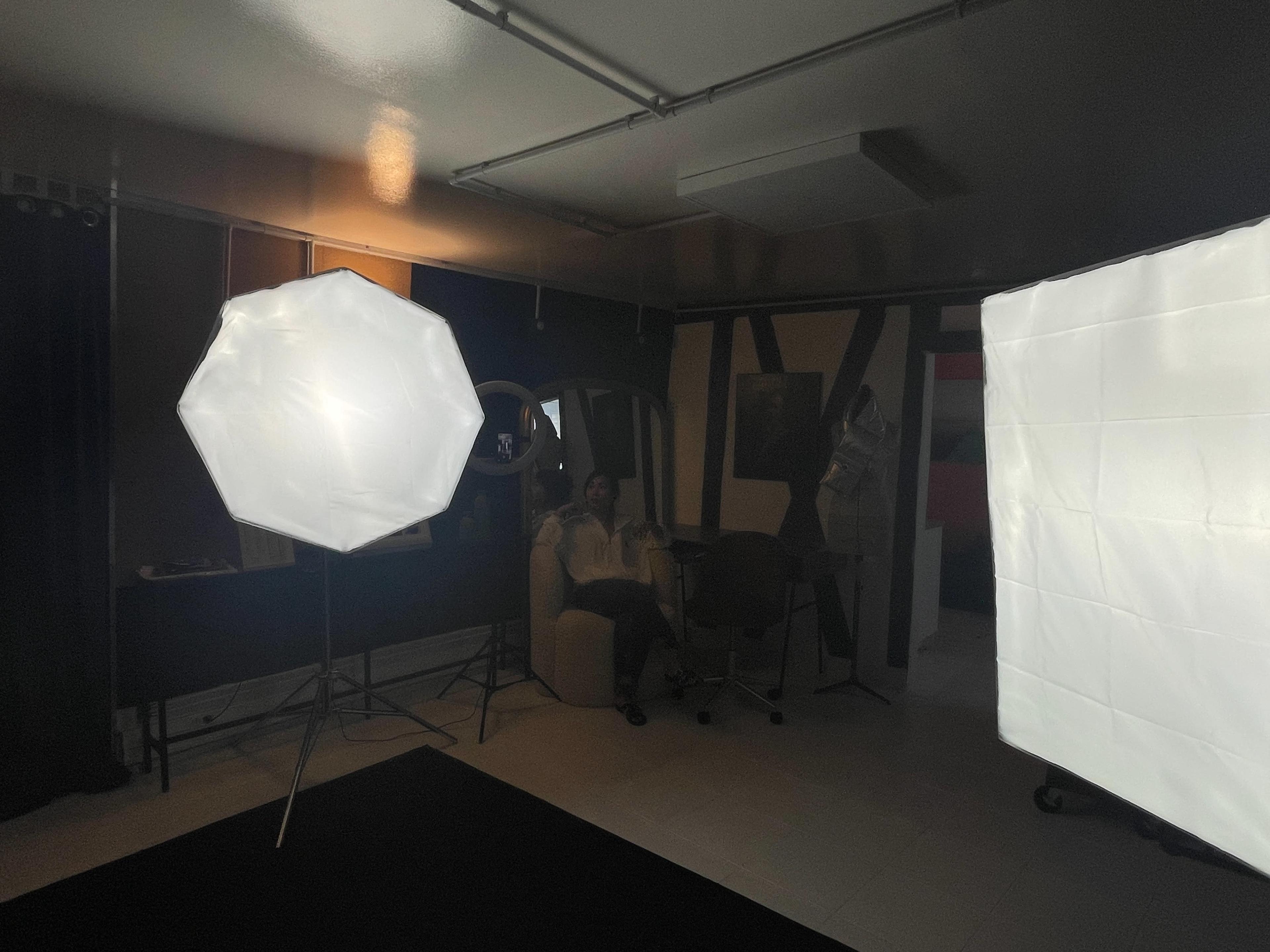 The image shows a dimly lit studio with two large softbox lights illuminating a woman seated in a chair against a backdrop of black curtains and various art pieces on the walls.