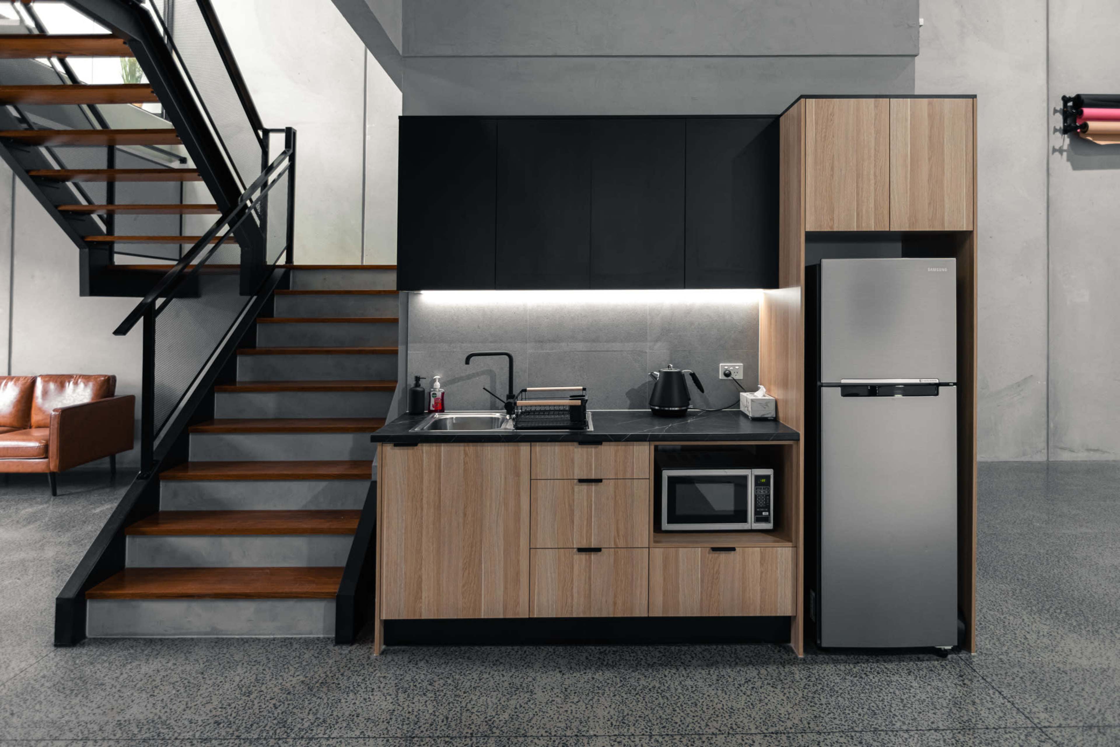 A modern kitchen with a black and wood finish, equipped with a refrigerator, microwave, kettle, and a staircase leading to an upper level.