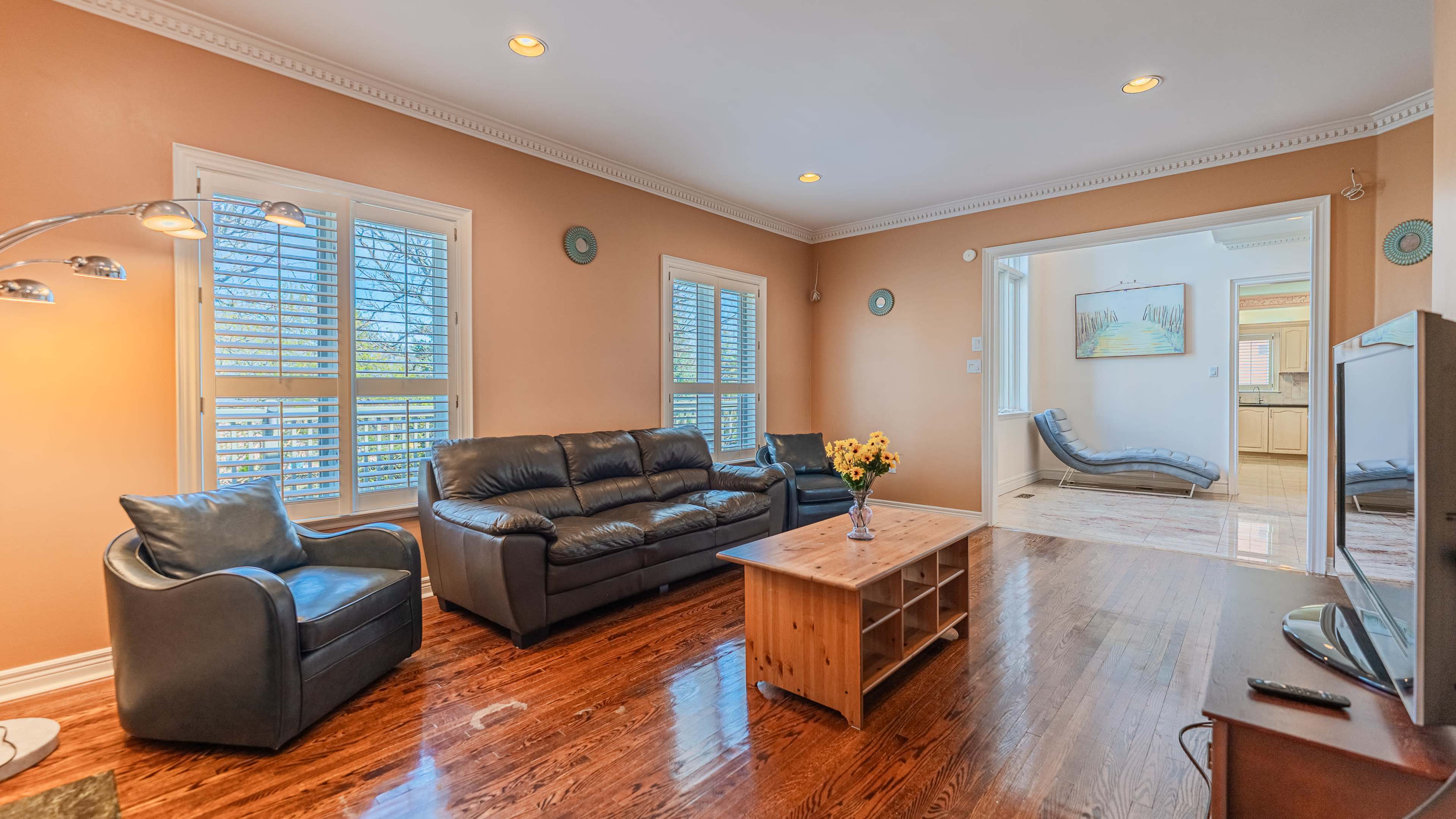 A living room features a dark leather sofa, a wooden coffee table, and light-colored walls with windows allowing natural light.