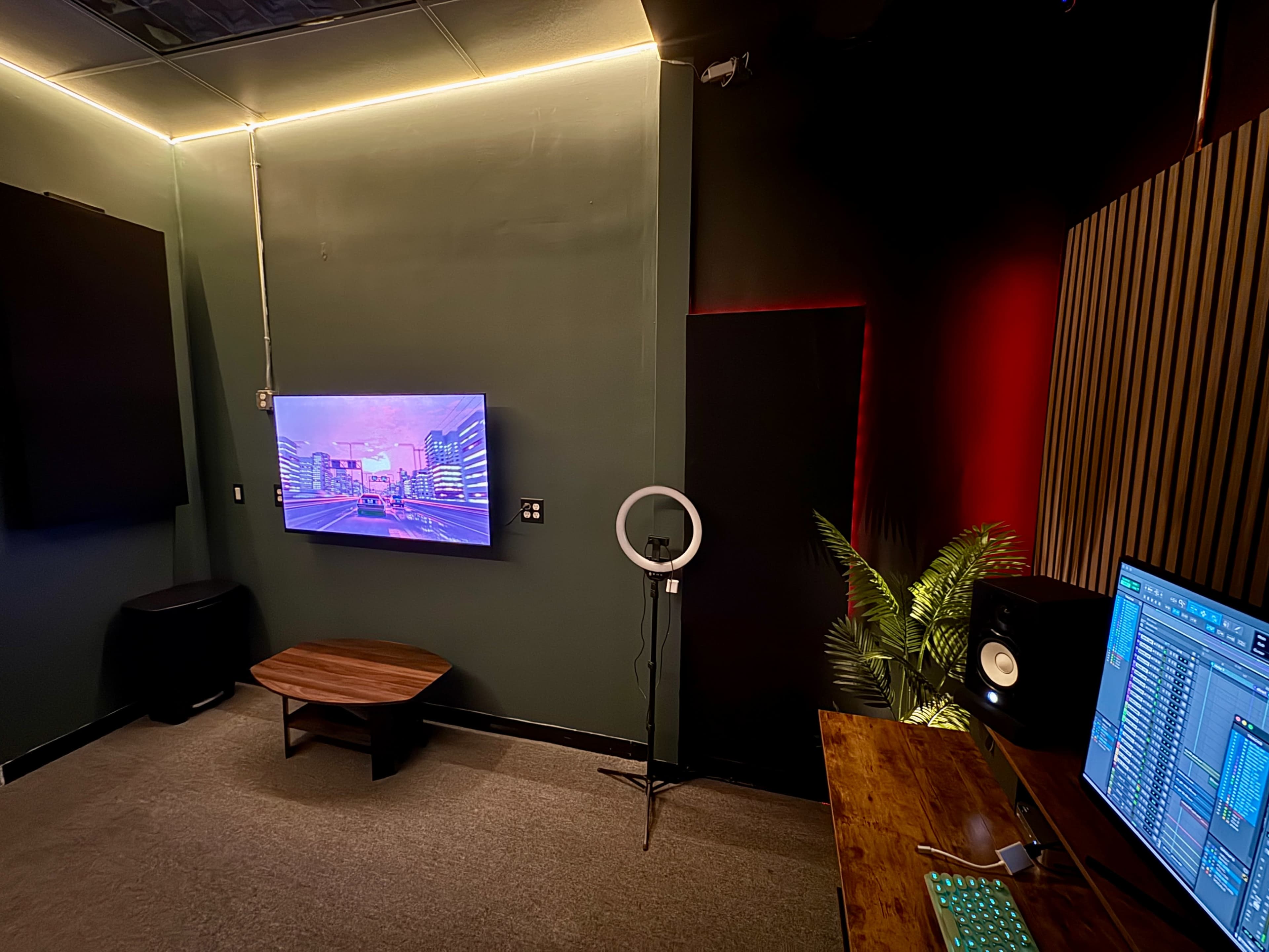 The image shows a corner of a music studio featuring a television screen displaying a cityscape, a ring light on a stand, a wooden table, a monitor with audio production software, and acoustic panels on the walls.