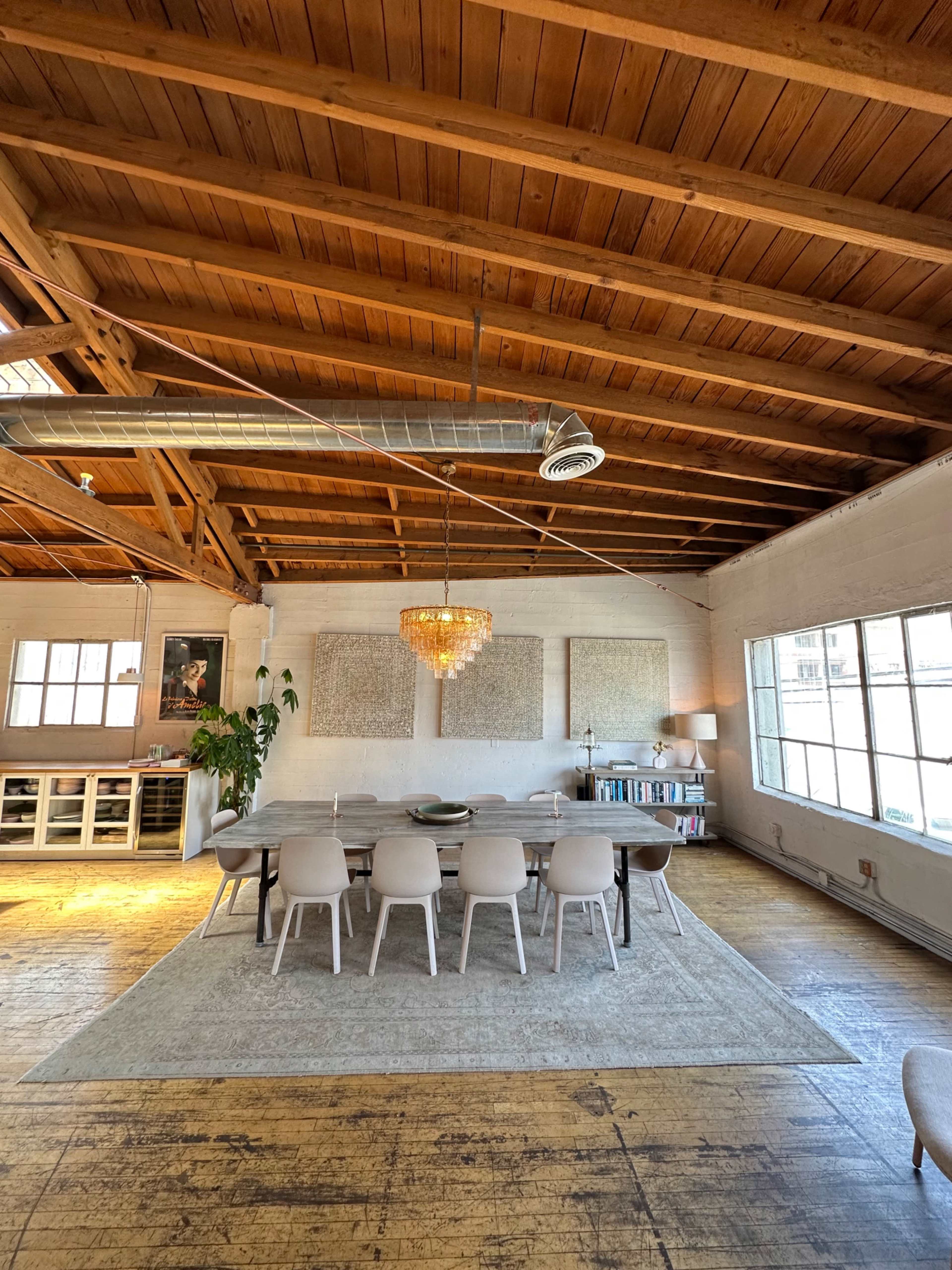 A spacious dining area features a long table surrounded by white chairs, with a chandelier hanging above and large windows letting in natural light.