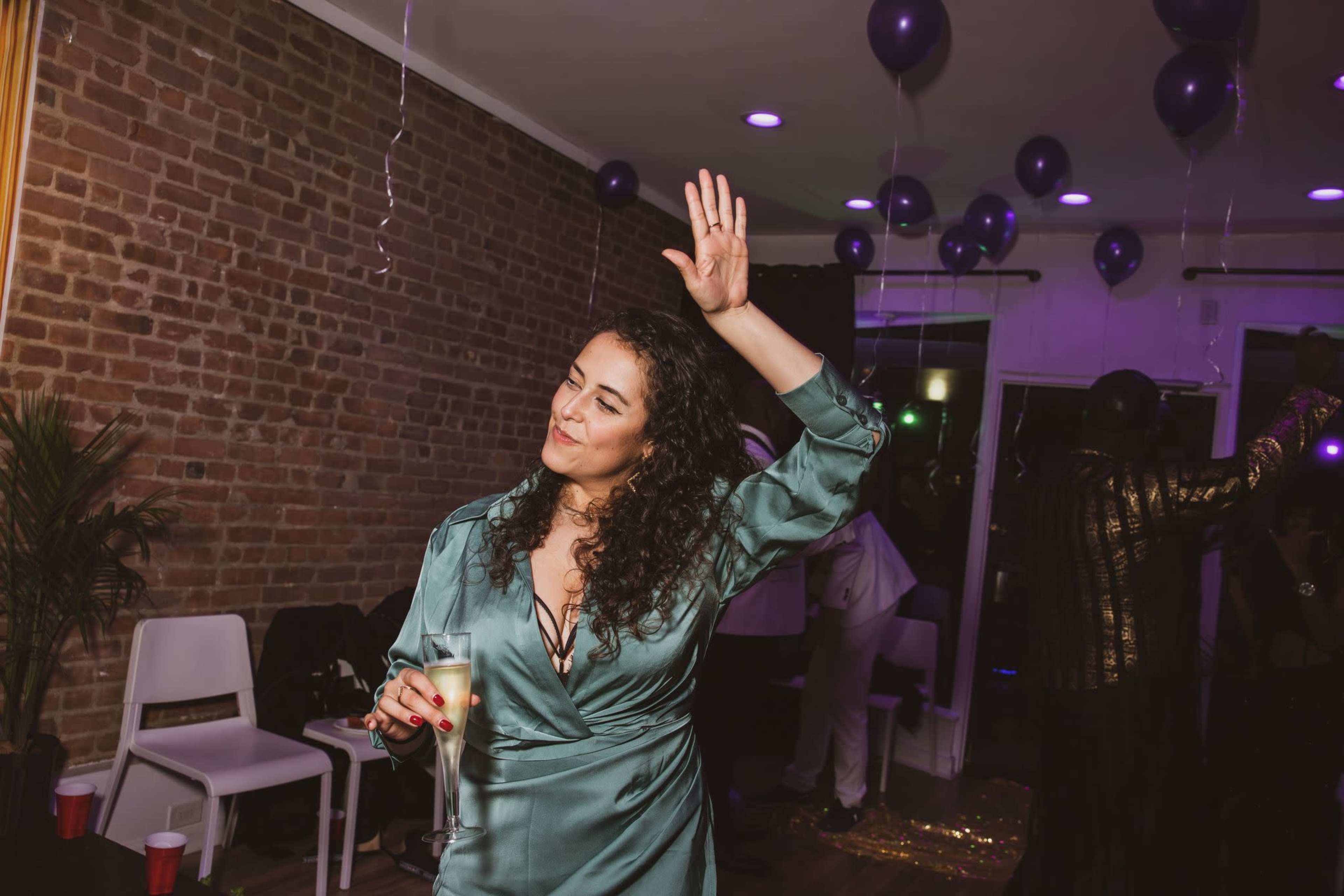 A woman in a green dress raises her hand while holding a drink at a festive indoor party with balloons and decorations.