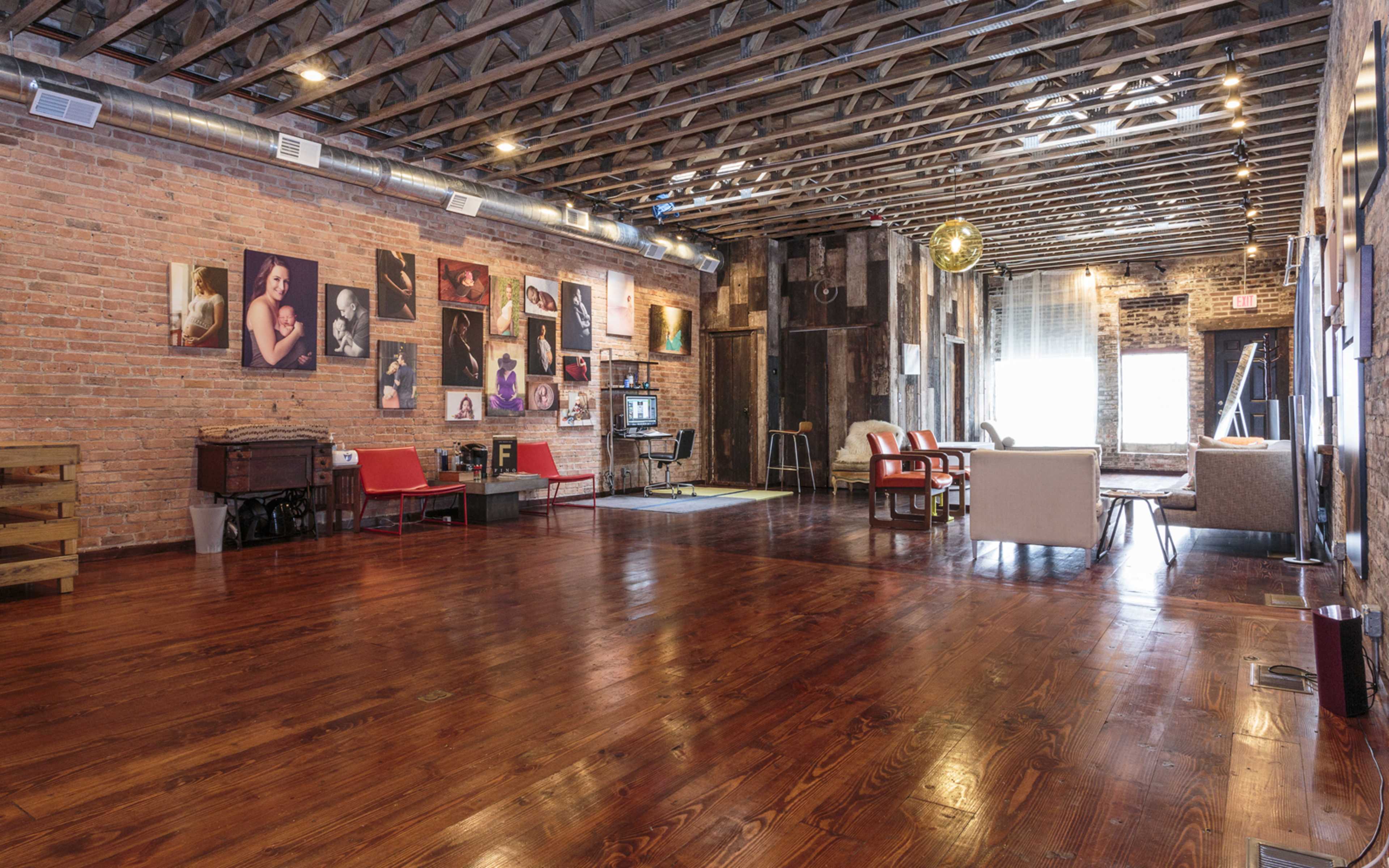 A spacious interior with exposed brick walls, wooden floors, and various seating arrangements, including red chairs and sofas, complemented by framed photographs on the walls.