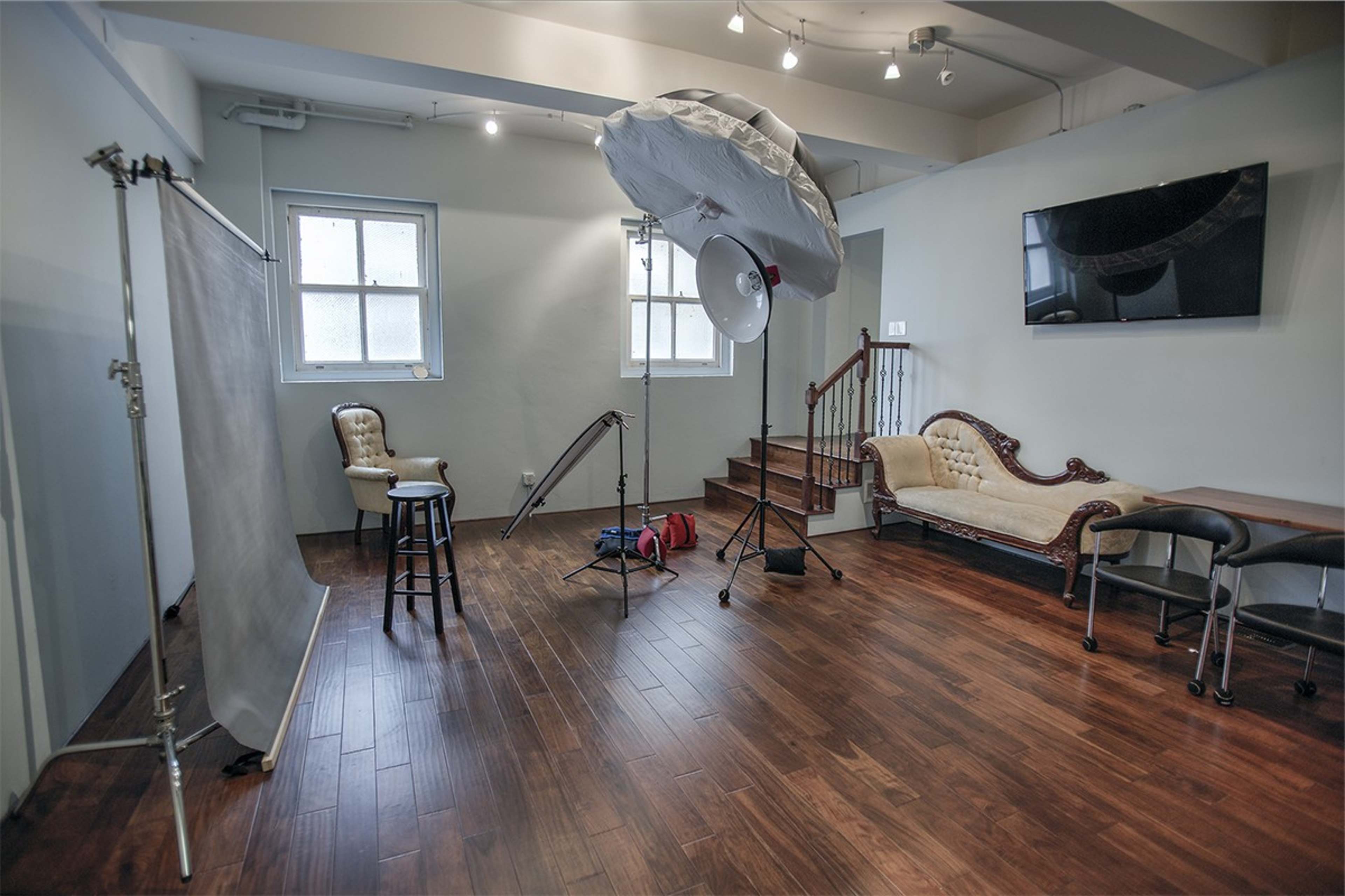 A photography studio with a backdrop, lighting equipment, a stool, and a Victorian-style couch arranged in a wooden-floored space.