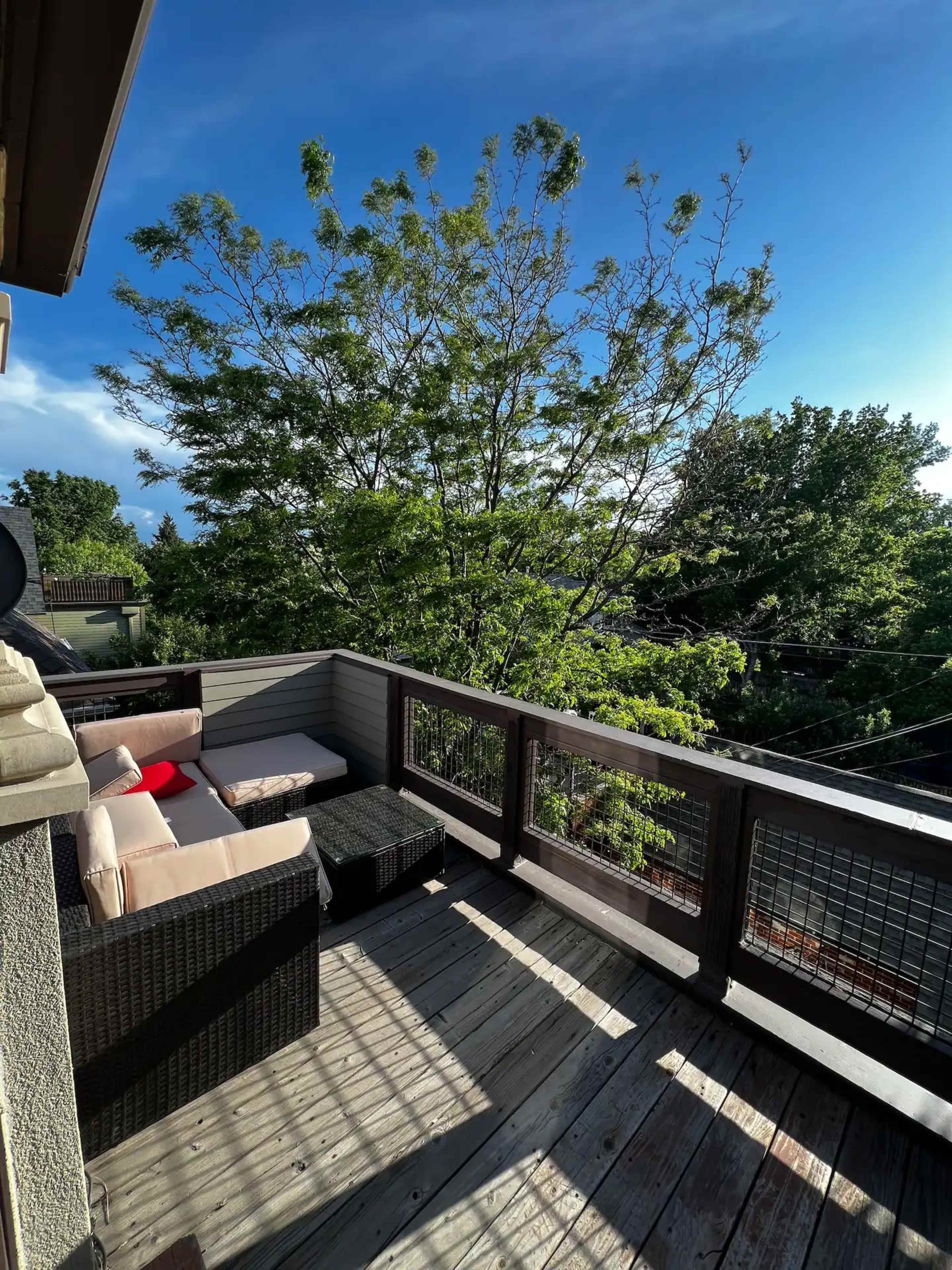A wooden balcony features a seating area with a light-colored sofa and a black coffee table, surrounded by trees and blue sky.