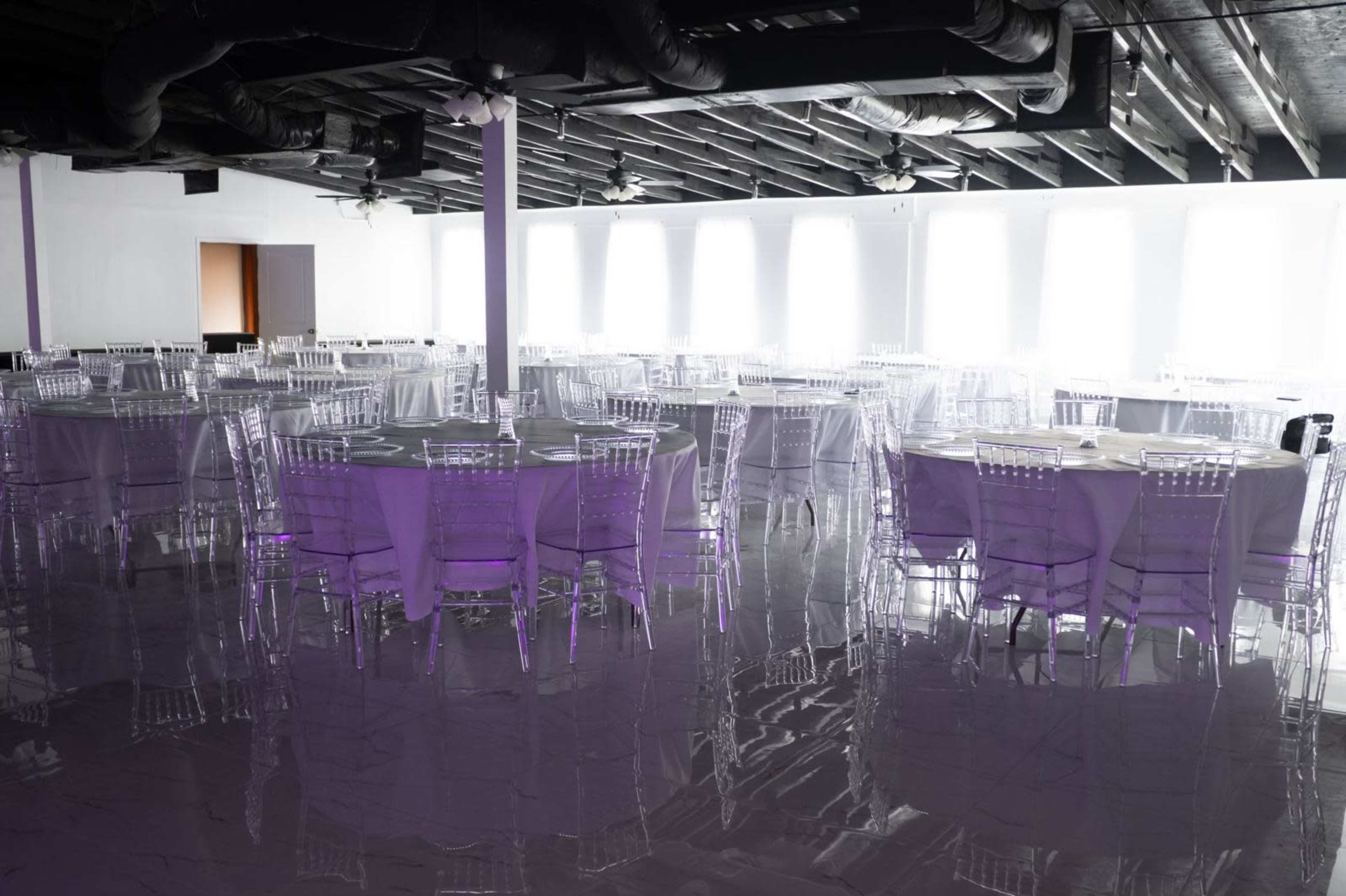 A spacious event venue features several round tables with clear chairs, set against a backdrop of large windows.