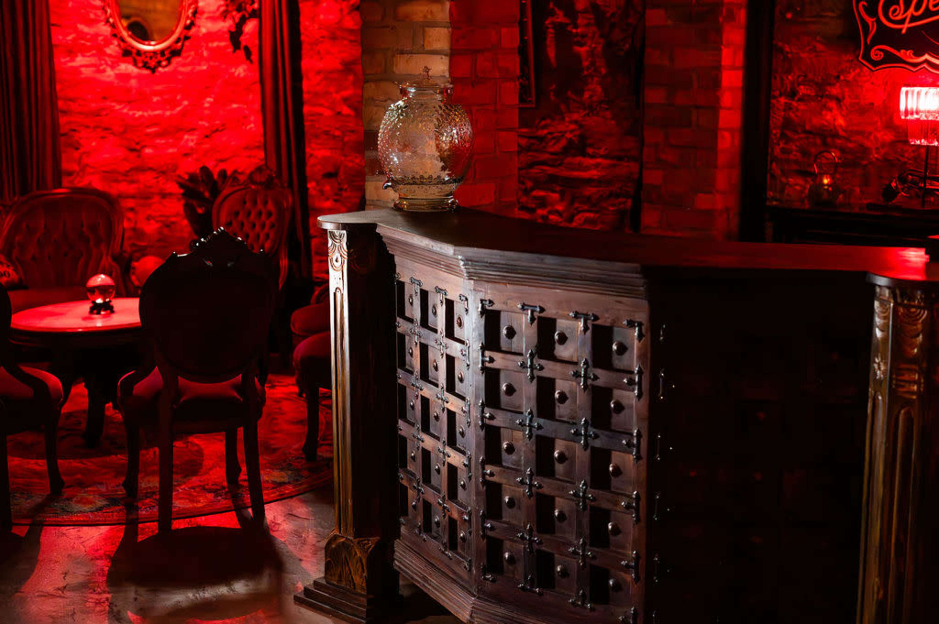 The image shows a dimly lit bar area with a wooden counter featuring several small drawers, surrounded by plush seating and red-lit decor.