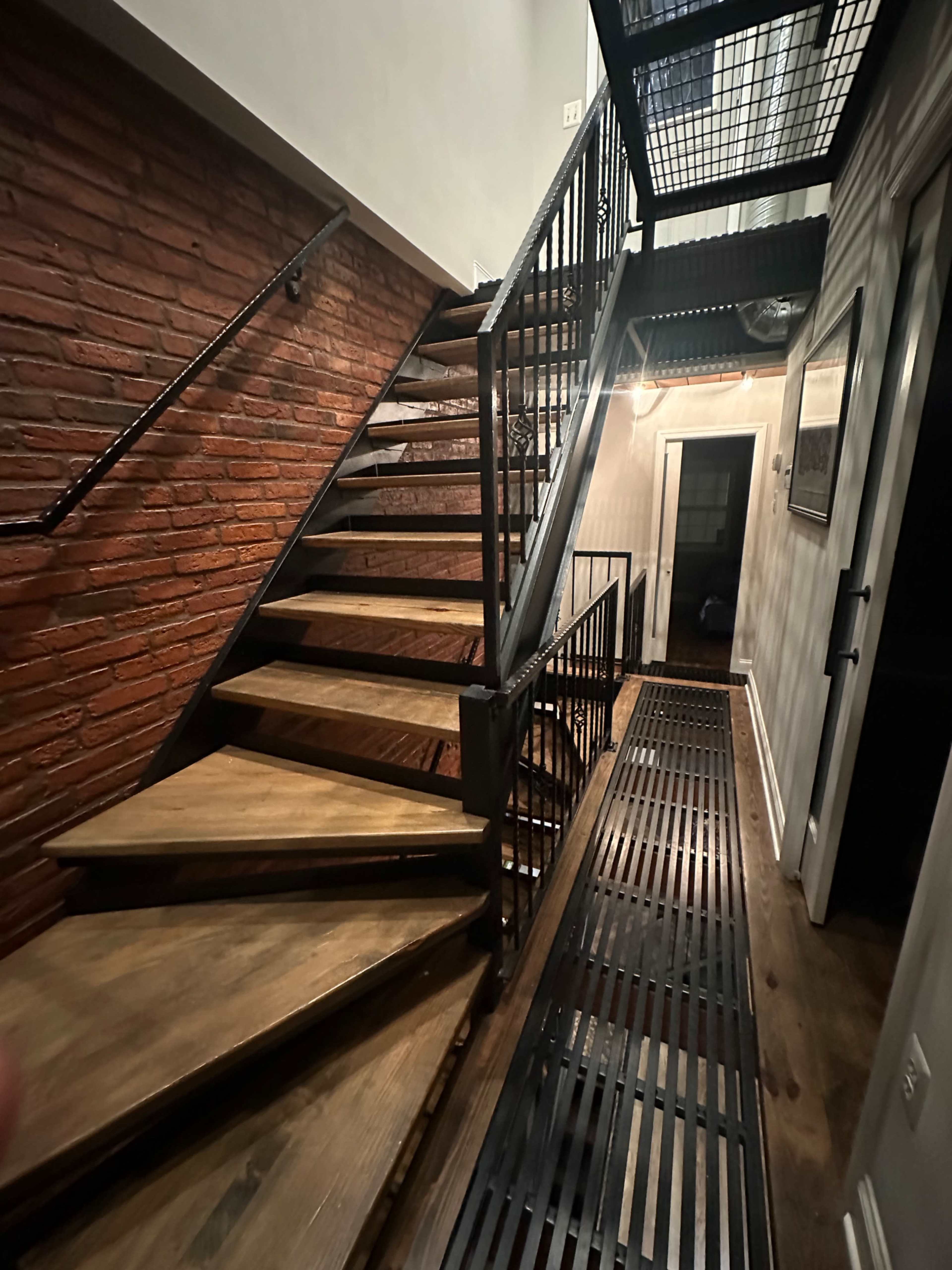 The image shows a metal and wooden staircase leading to a corridor with a brick wall and industrial-style railings.