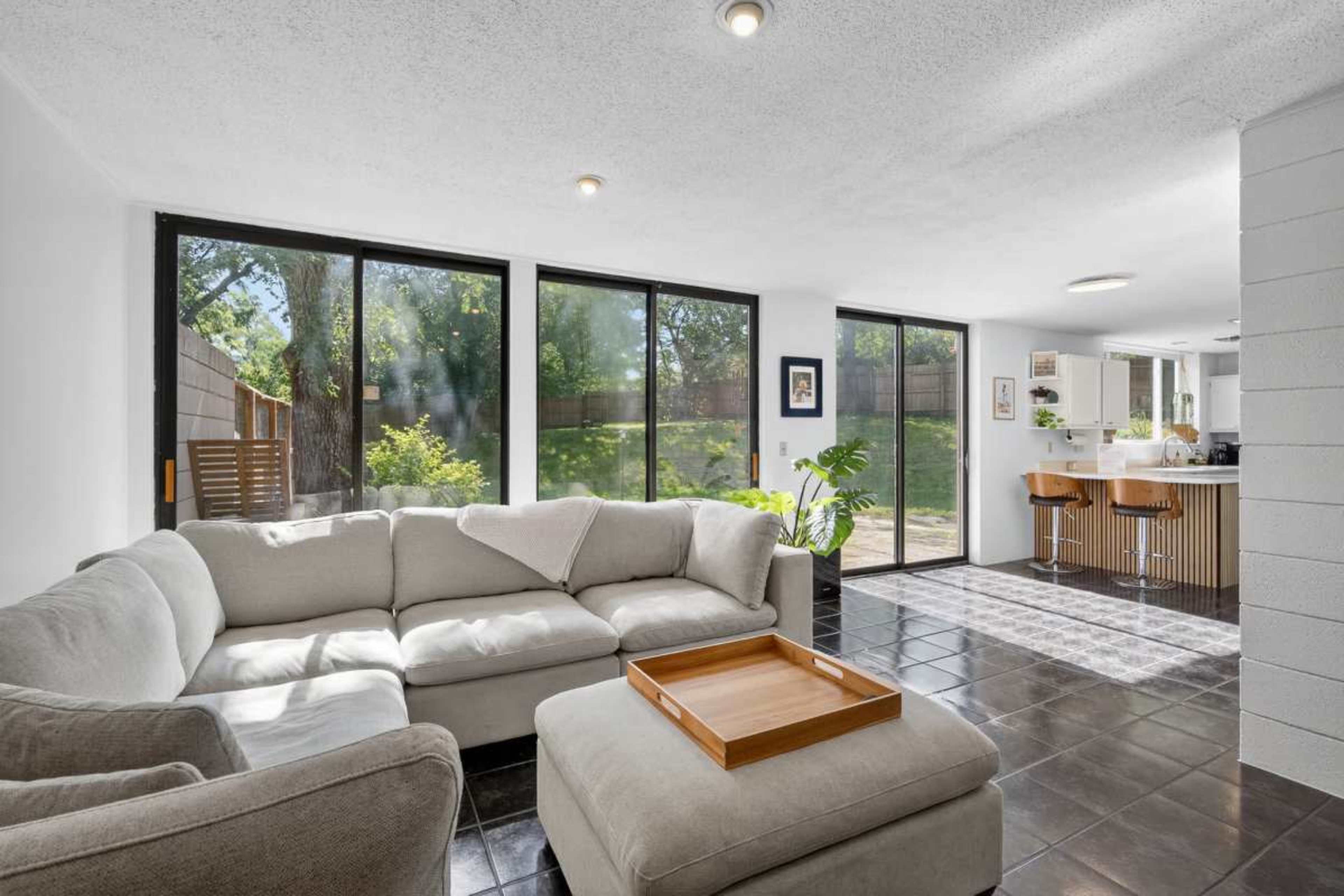 A modern living room features large windows with a view of a garden, a light-colored sectional sofa, and a tile floor.