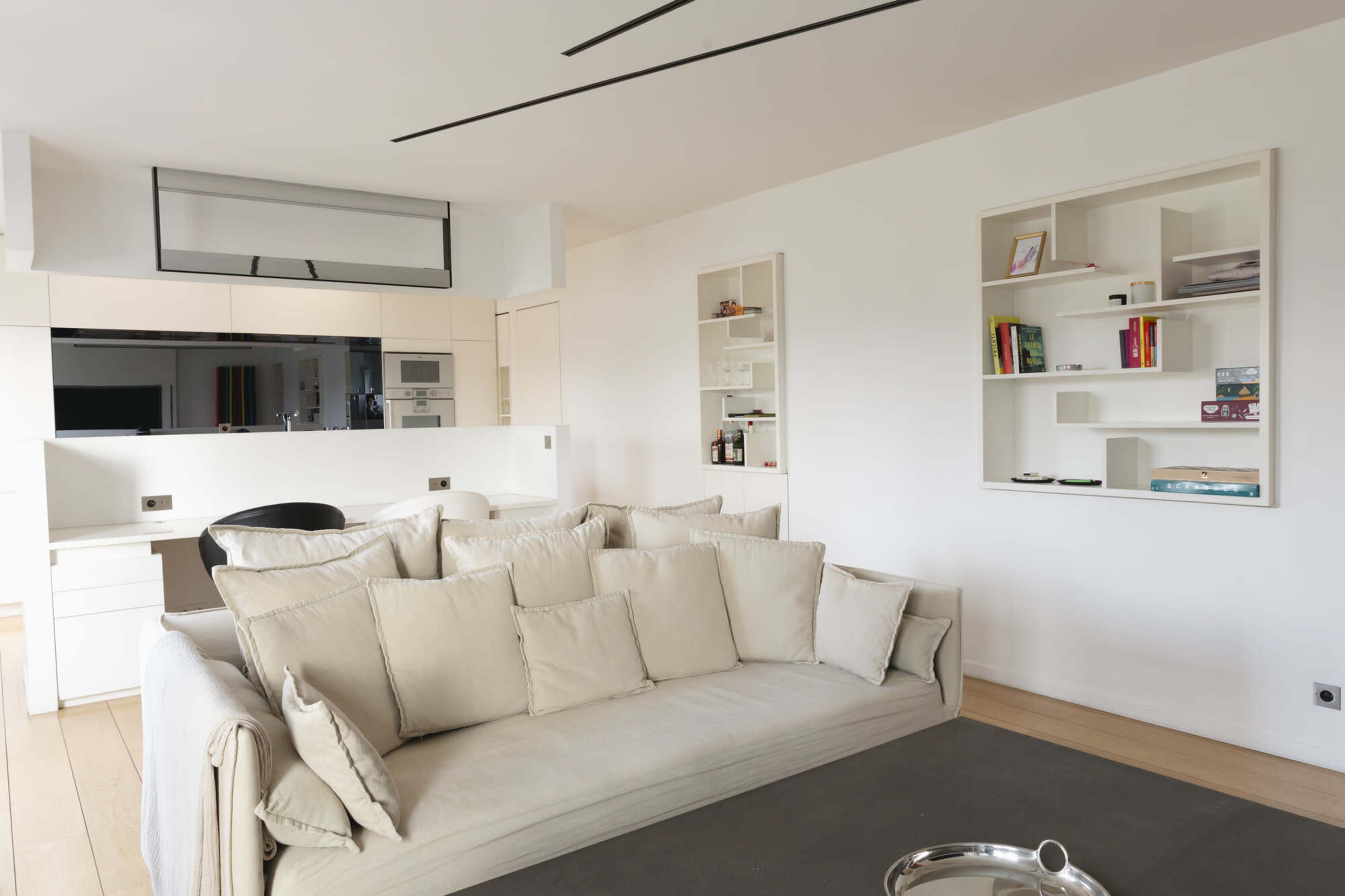 The image shows a modern living room with a large beige sofa, a dark area rug, and wall-mounted shelves displaying various items.