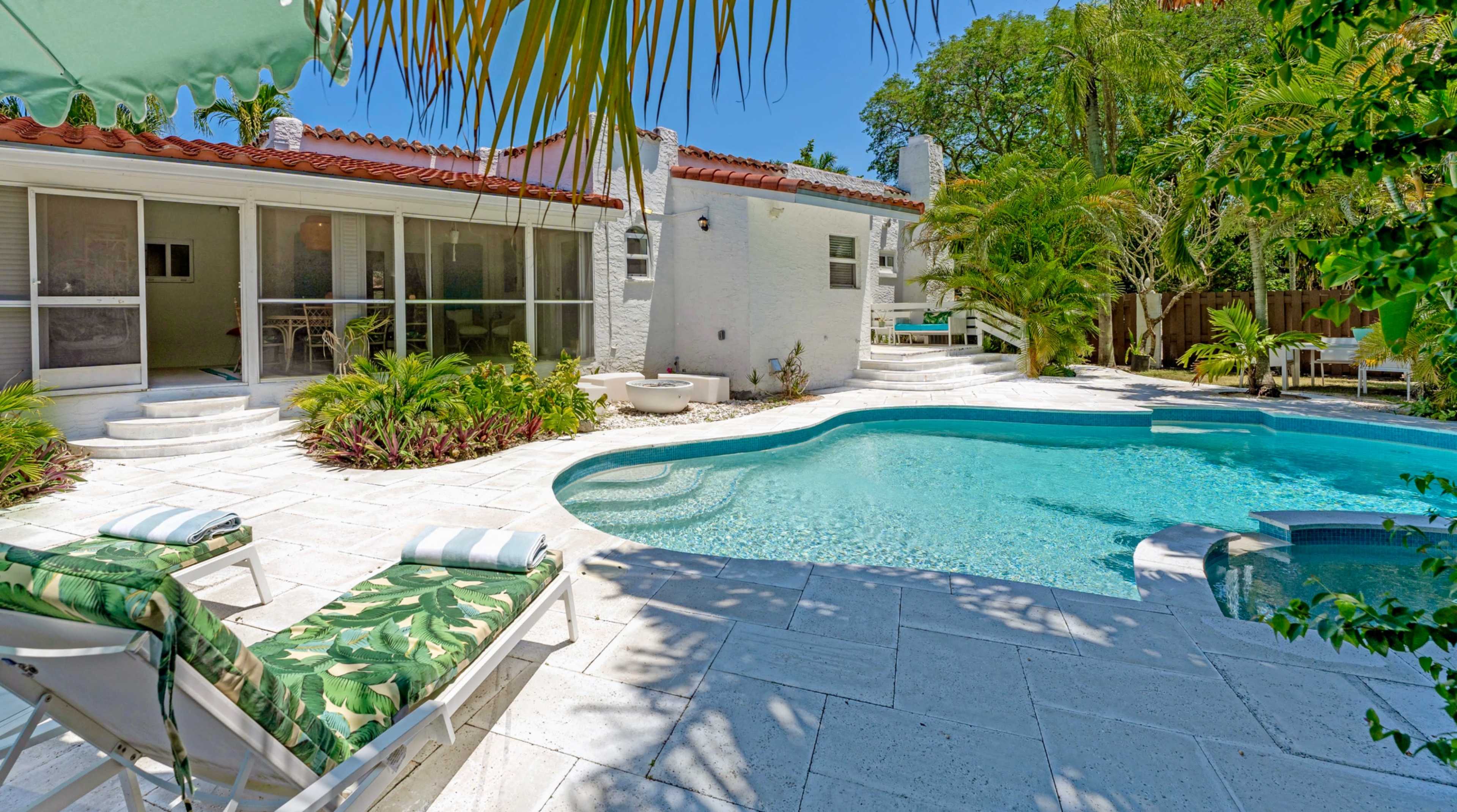 The image shows a bright outdoor area featuring a swimming pool surrounded by tropical landscaping and lounge chairs.