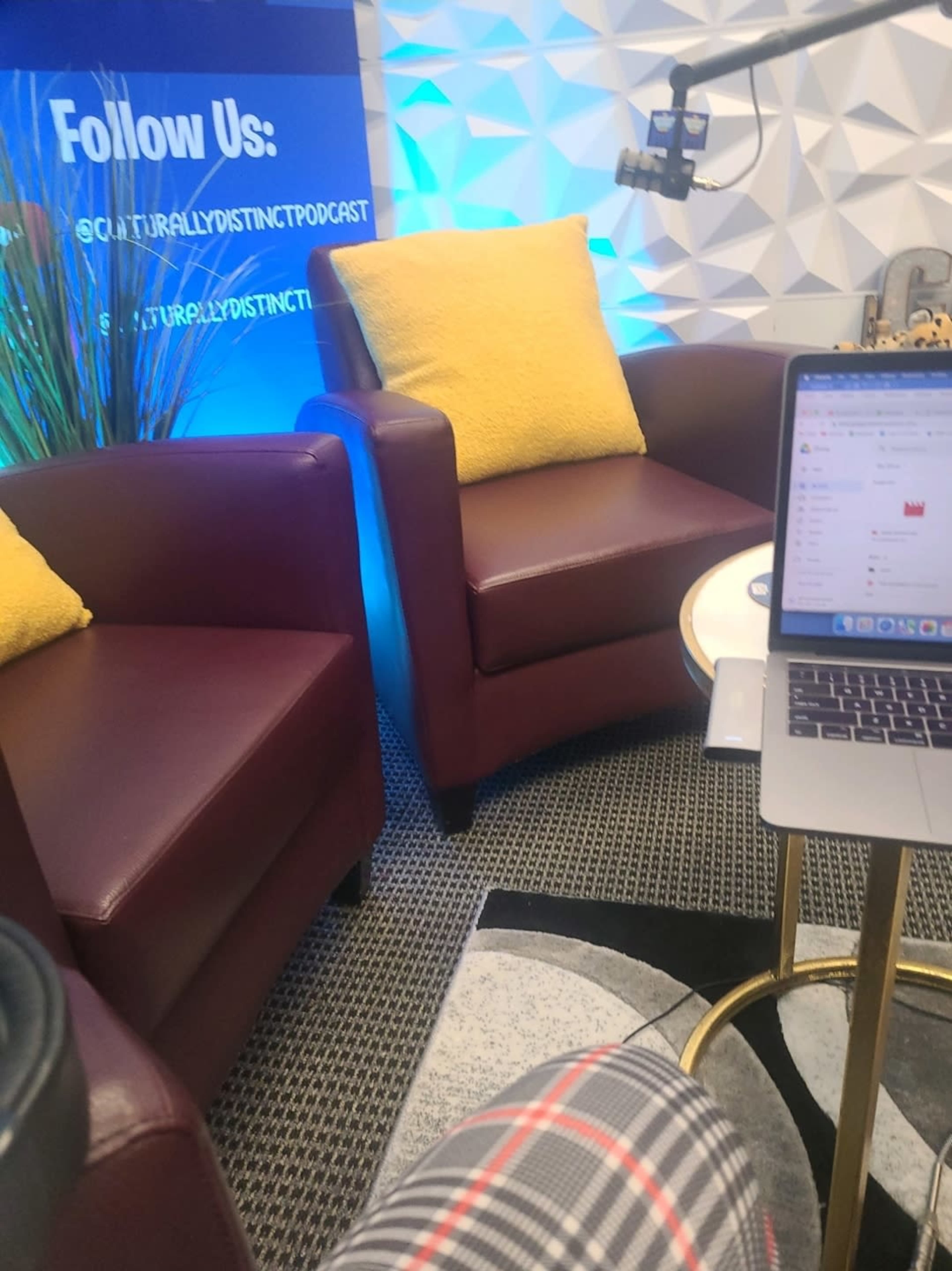 The image shows two maroon armchairs with yellow cushions, a small round table, and a laptop on a patterned rug in a podcast recording space.