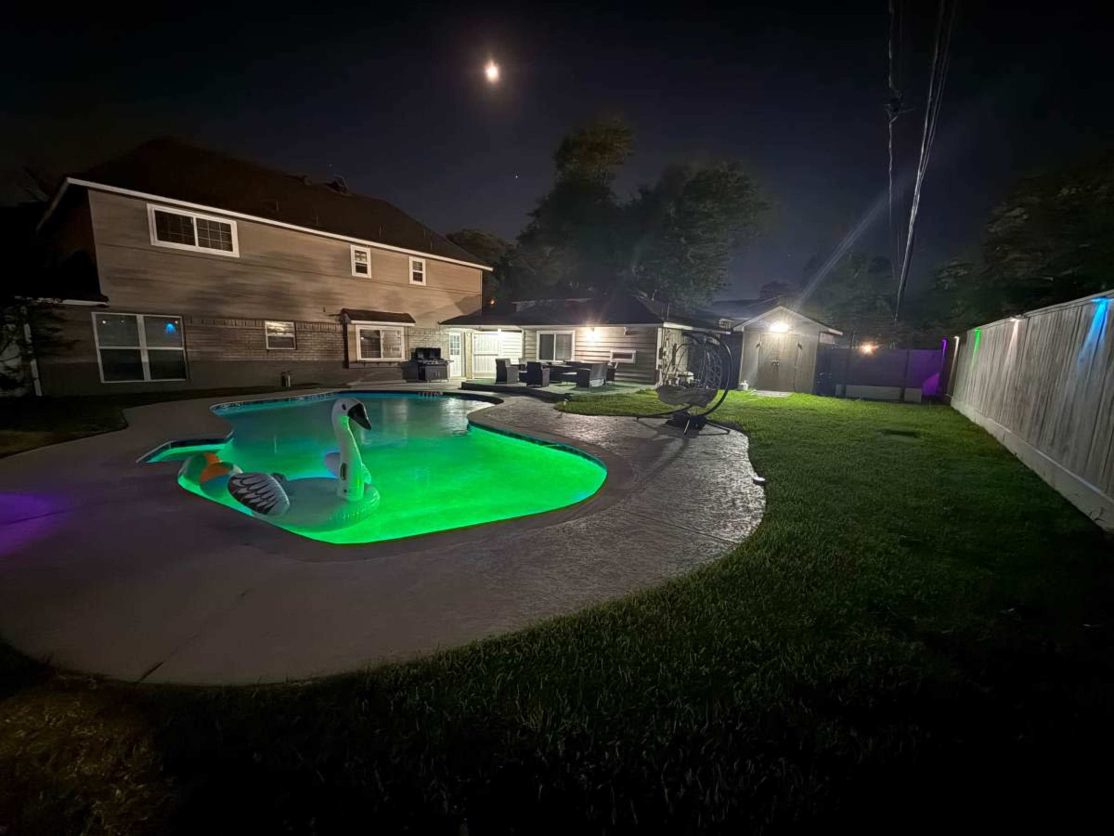 The image shows a backyard at night featuring a swimming pool illuminated with green lights, a swan float, and a house visible in the background.